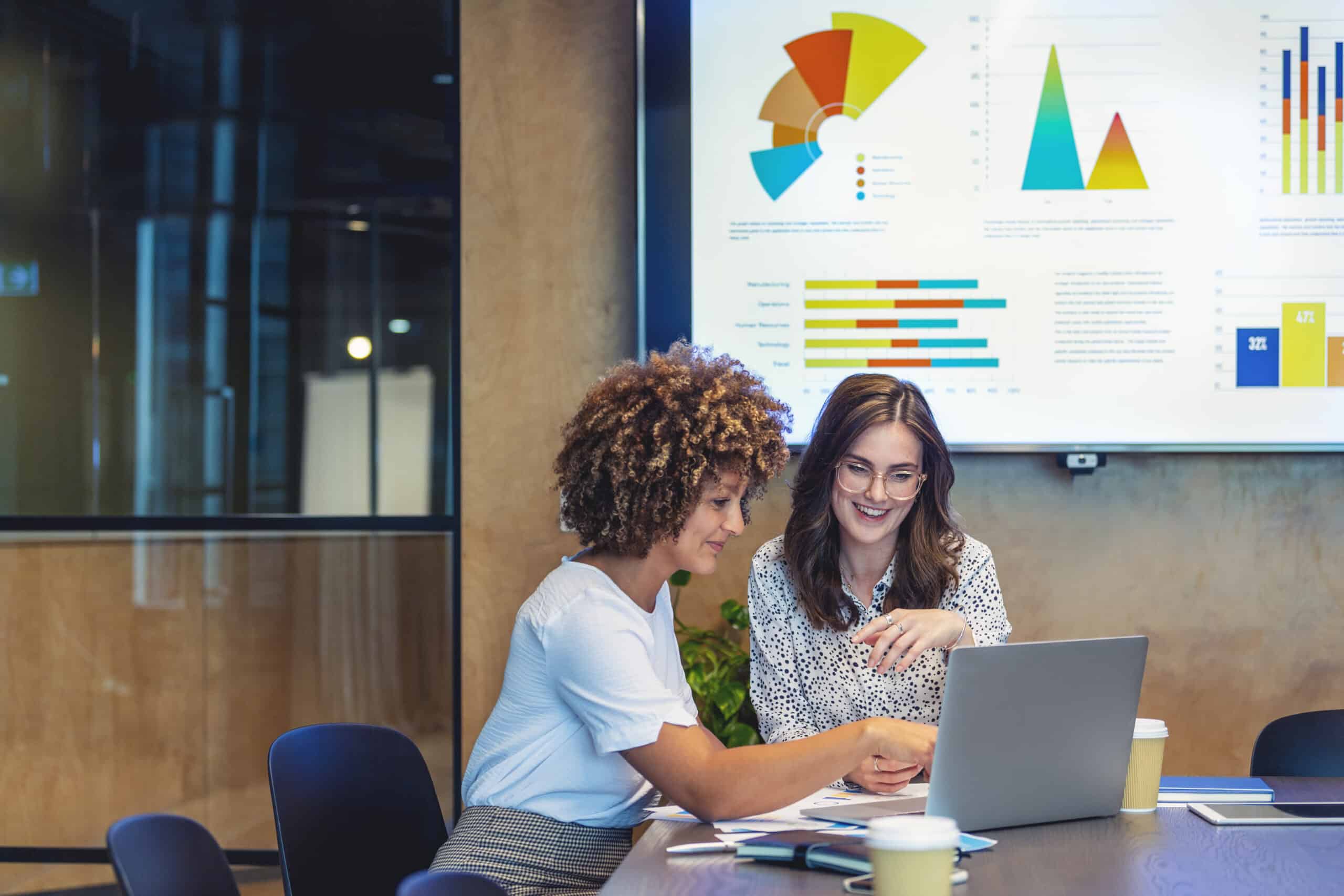 Two women discussing real estate fair market value analysis in a meeting room with charts behind them.