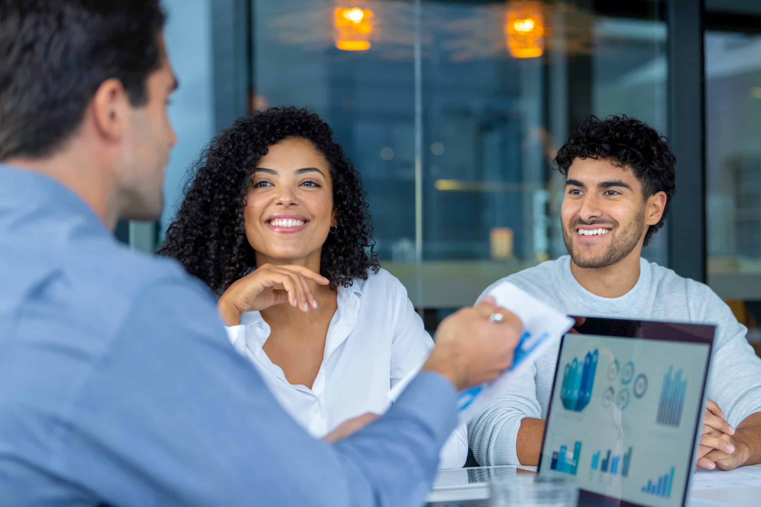 Three people have a business meeting, smiling, with a laptop displaying charts.