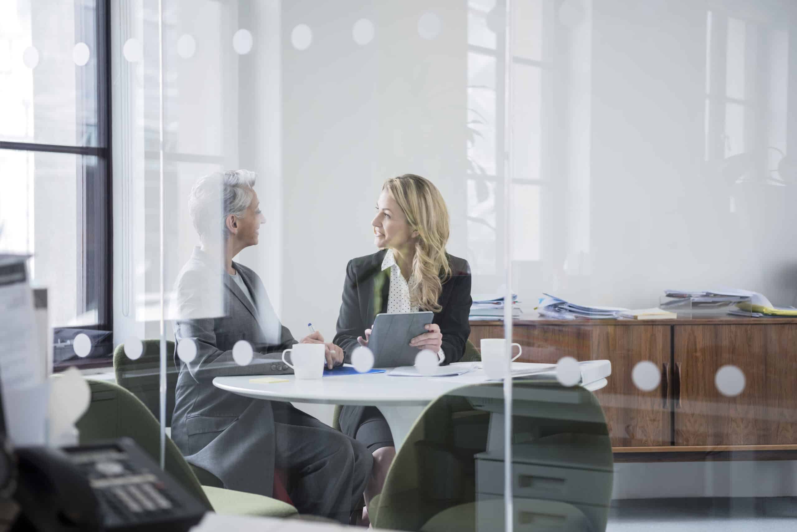 Two women in business attire discuss Eminent Domain Consulting at a round office table.