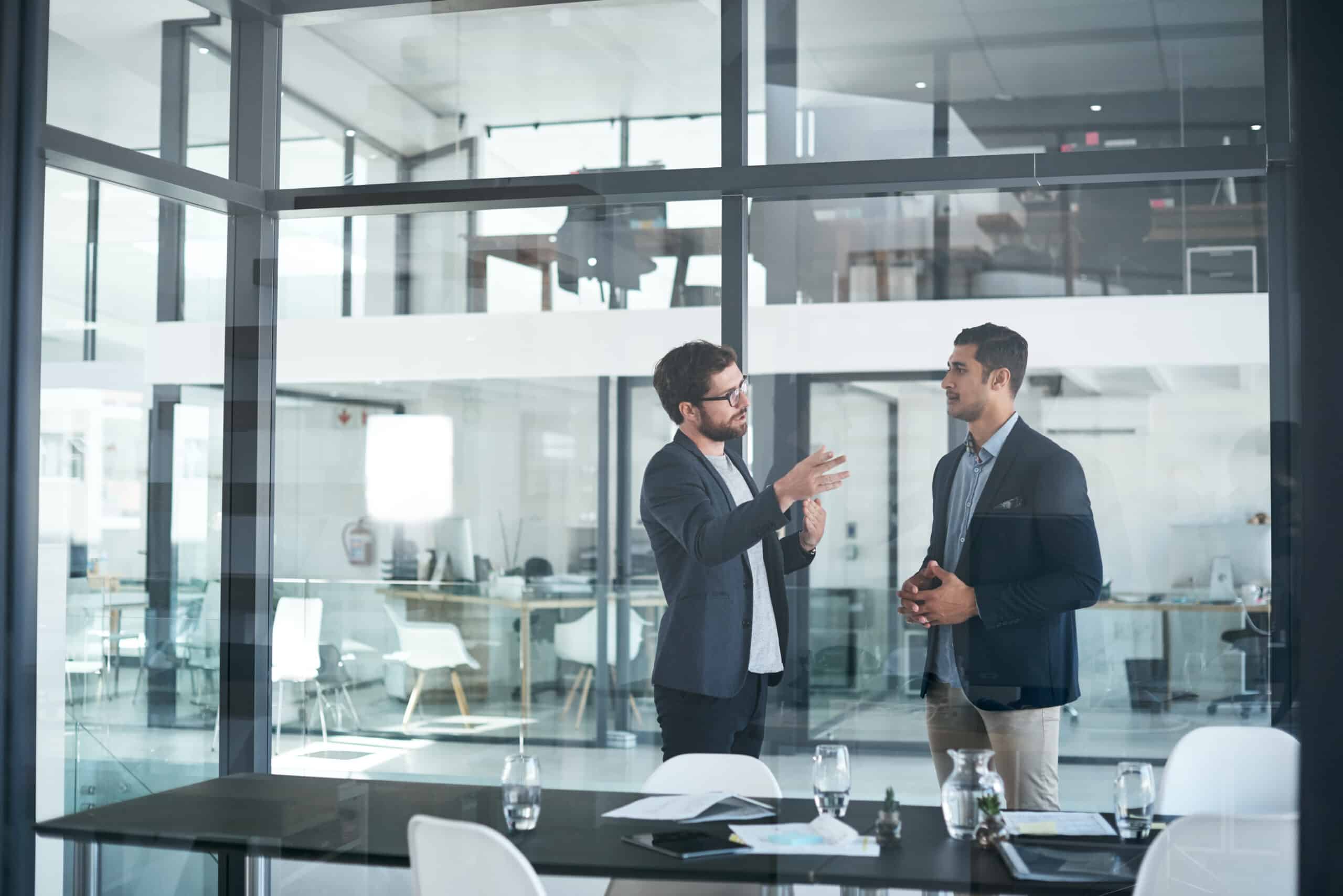 Two men in business attire discuss Eminent Domain consulting in a modern glass-walled office.
