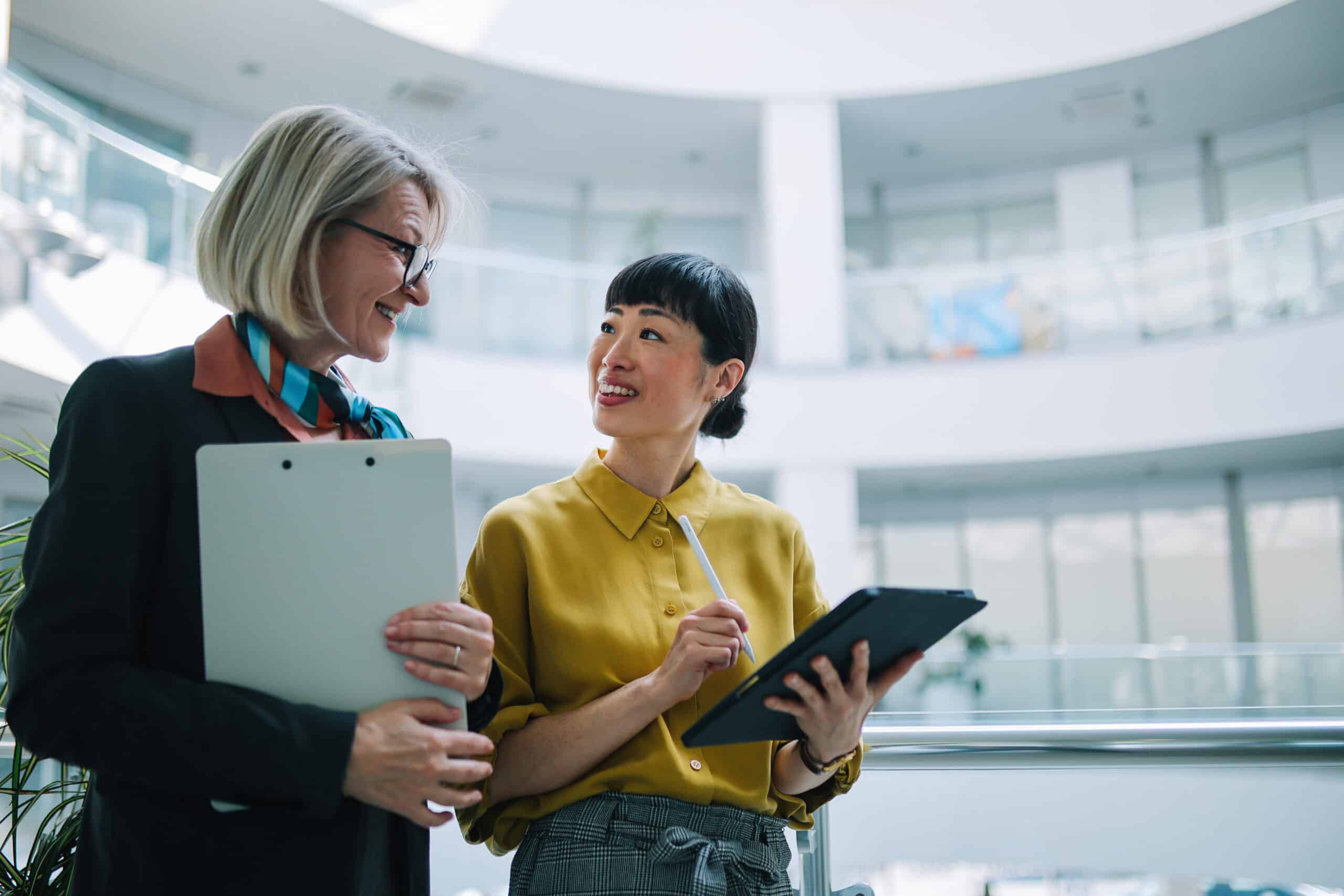 Two women in business attire discuss IT Modernization Consulting while holding a clipboard and tablet.