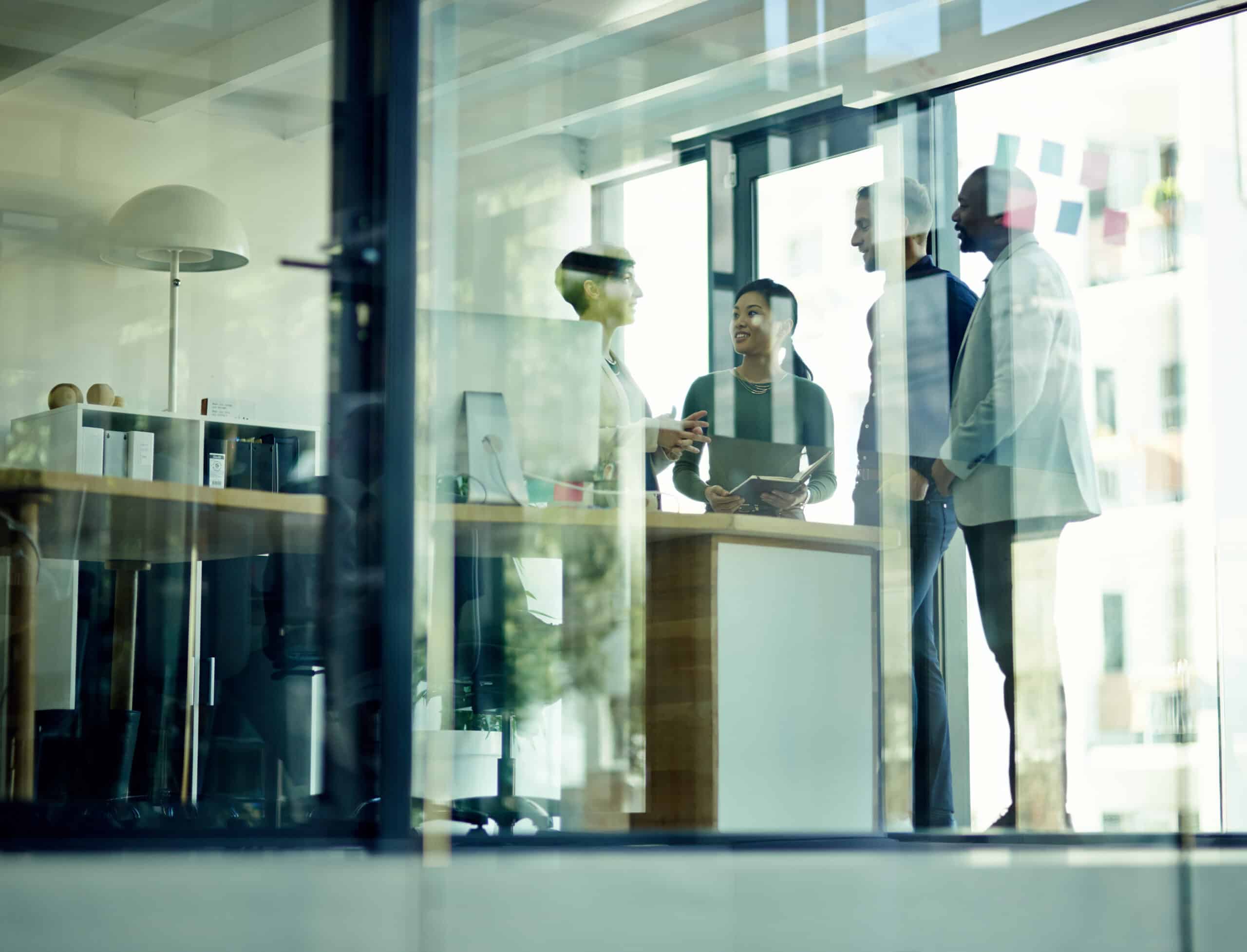 Four people discuss with AI consultants in a modern glass-walled office with desks and computers.