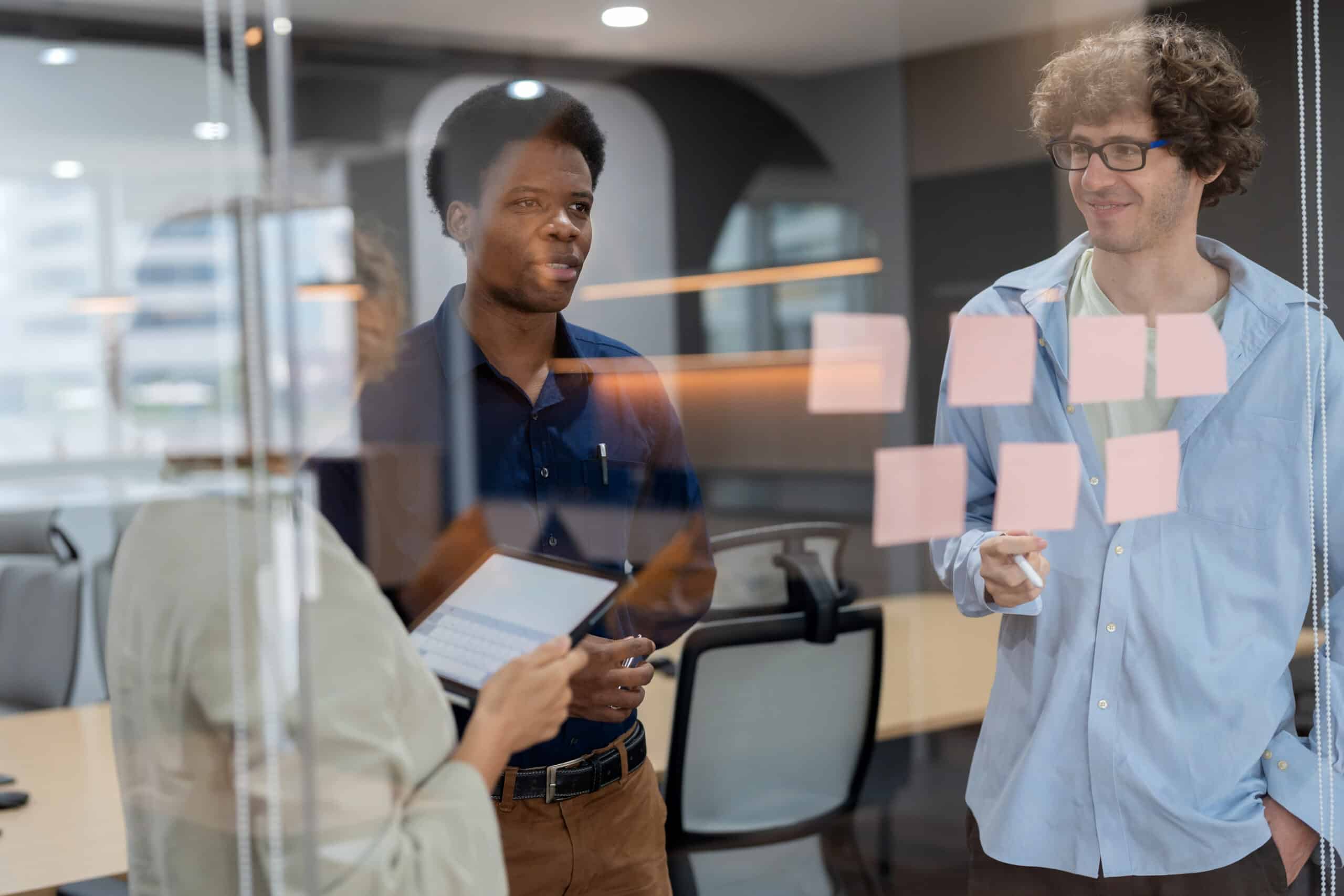 Head of product development standing in a meeting room, discussing ideas with a multiracial team while posting adhesive notes on the glass wall. The session focuses on analyzing market data, creating strategies, and driving business innovation.