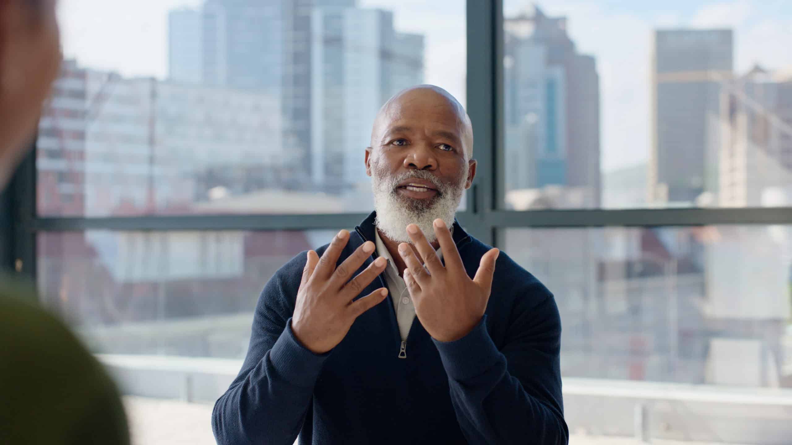 Middle-aged man talking with expressive hand gestures in a bright office with city views.