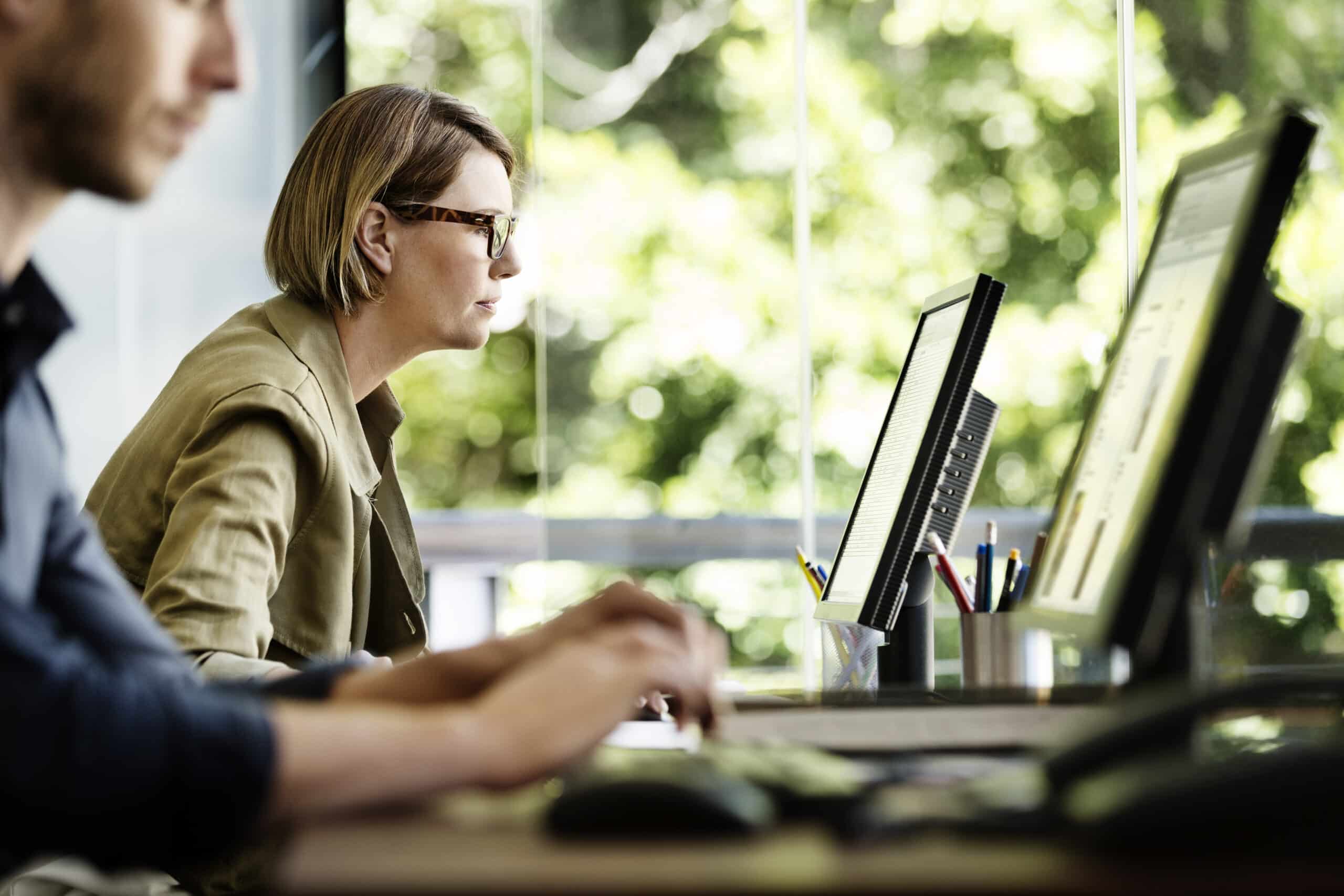Two people work on desktops in a smart building office with large windows and greenery outside.