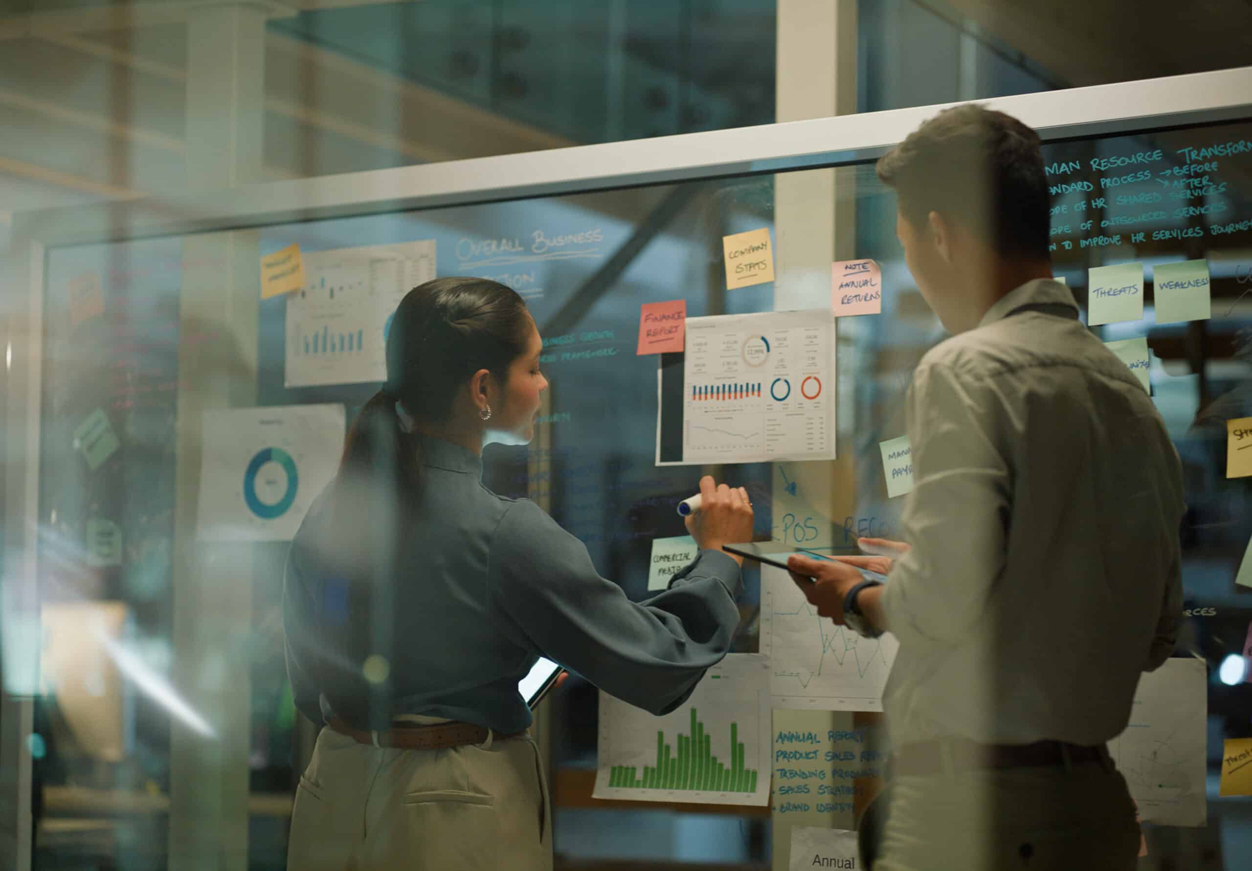 Two IoT consultants reviewing charts and graphs on a glass wall covered with notes and papers.