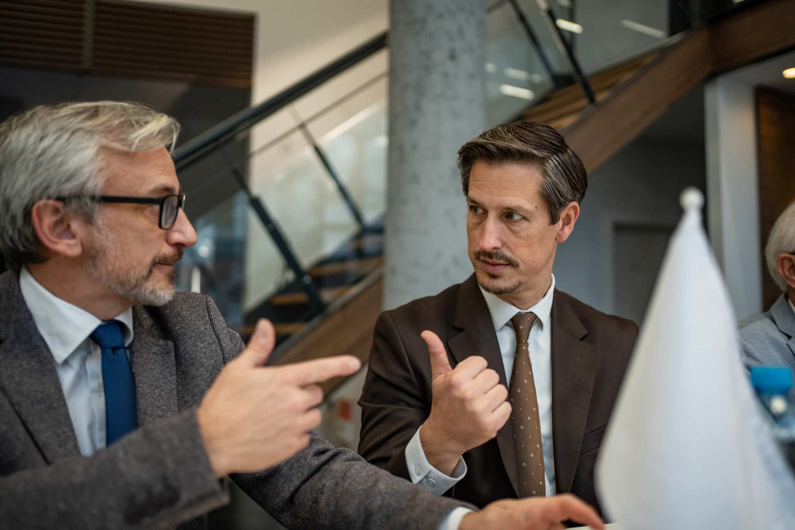 Two men in suits discuss CRE Workplace Strategy at a meeting table with a white flag in front.