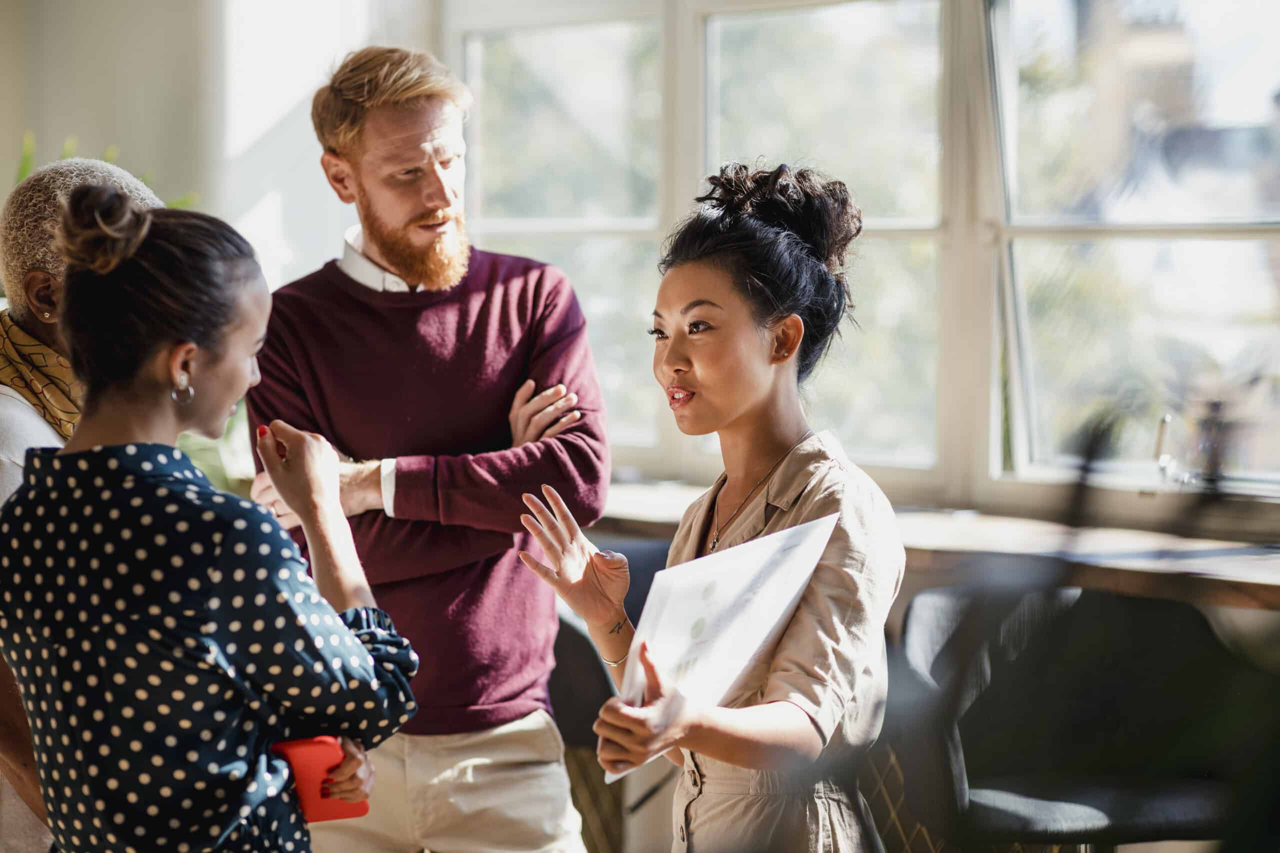 Three people stand in a sunlit office, having a discussion, one holding a laptop.