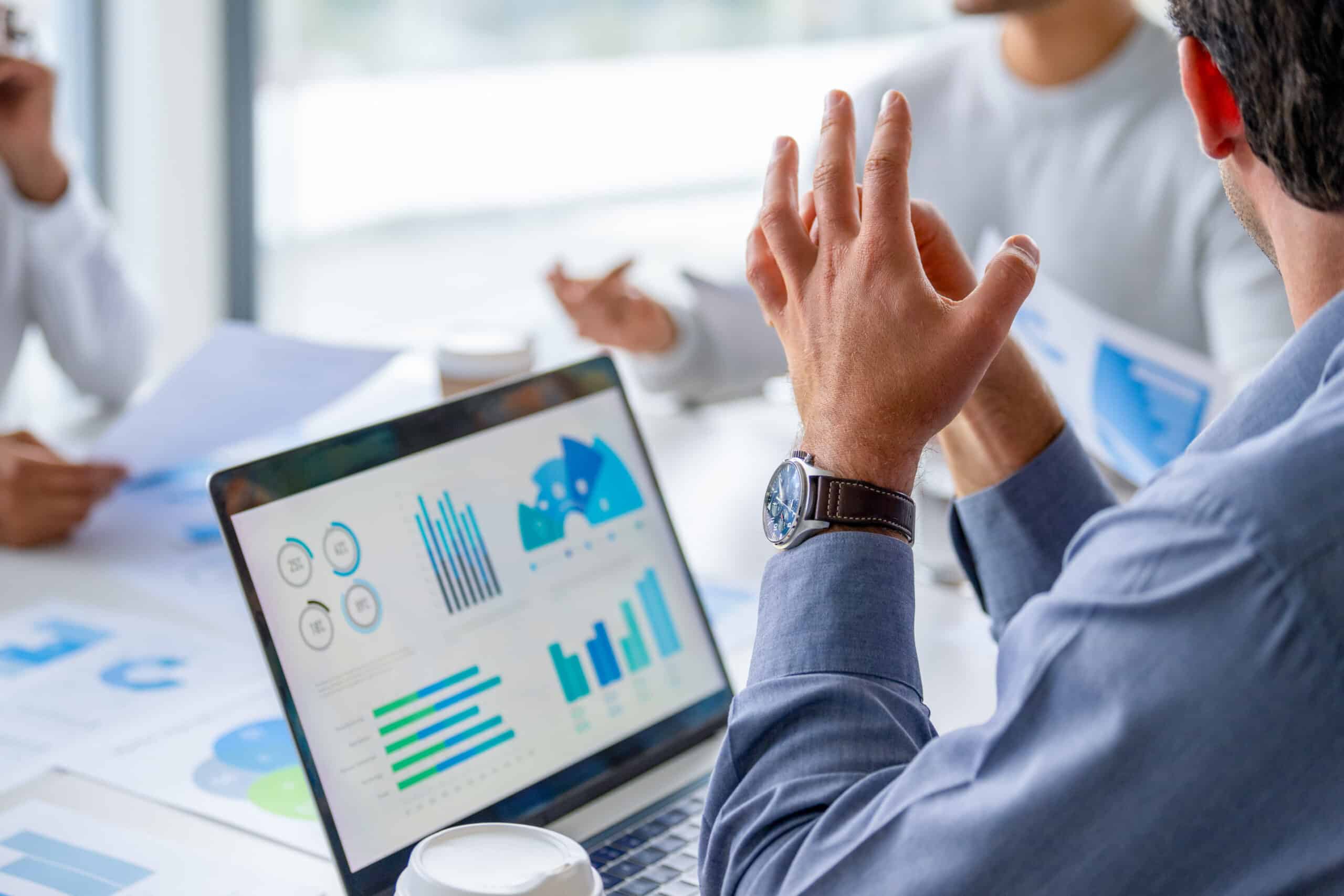 Close up of a laptop and document with data and finance charts and graphs. Group of business people in a meeting in then board room in the background.