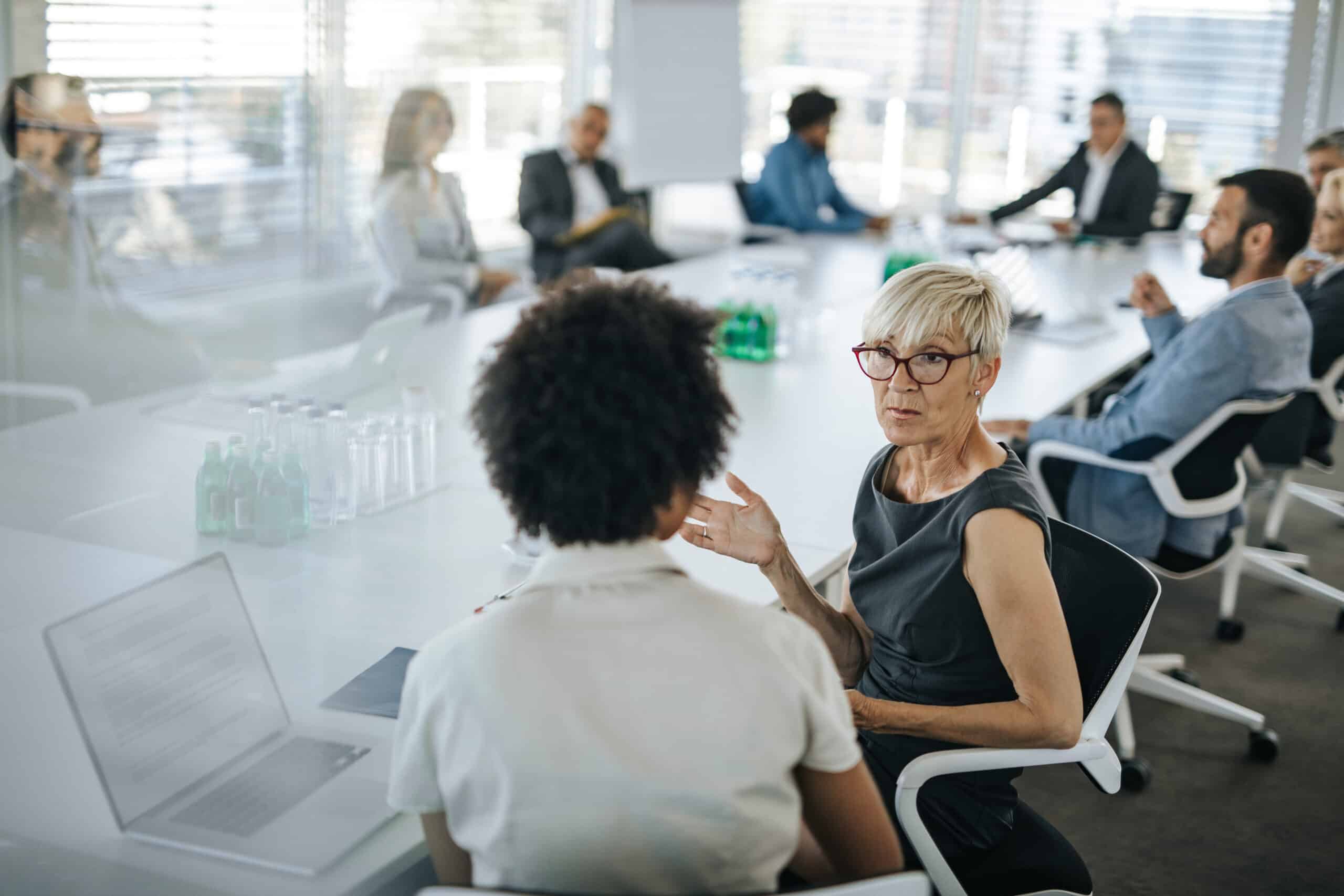 Two women discuss Eminent Domain Consulting during a meeting with several colleagues.