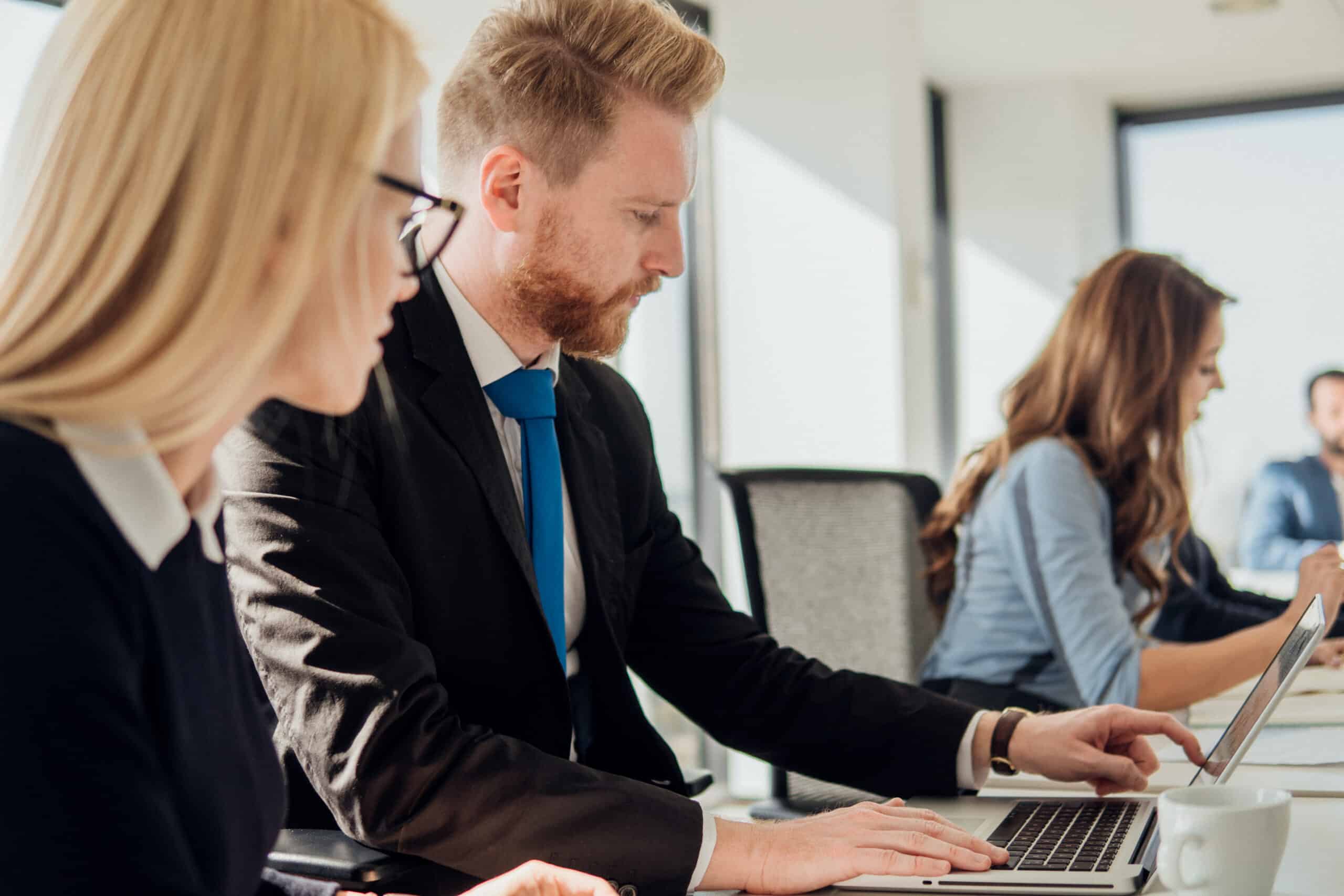 Two people in business attire work on laptops at a bright Smart Building Consulting office desk.