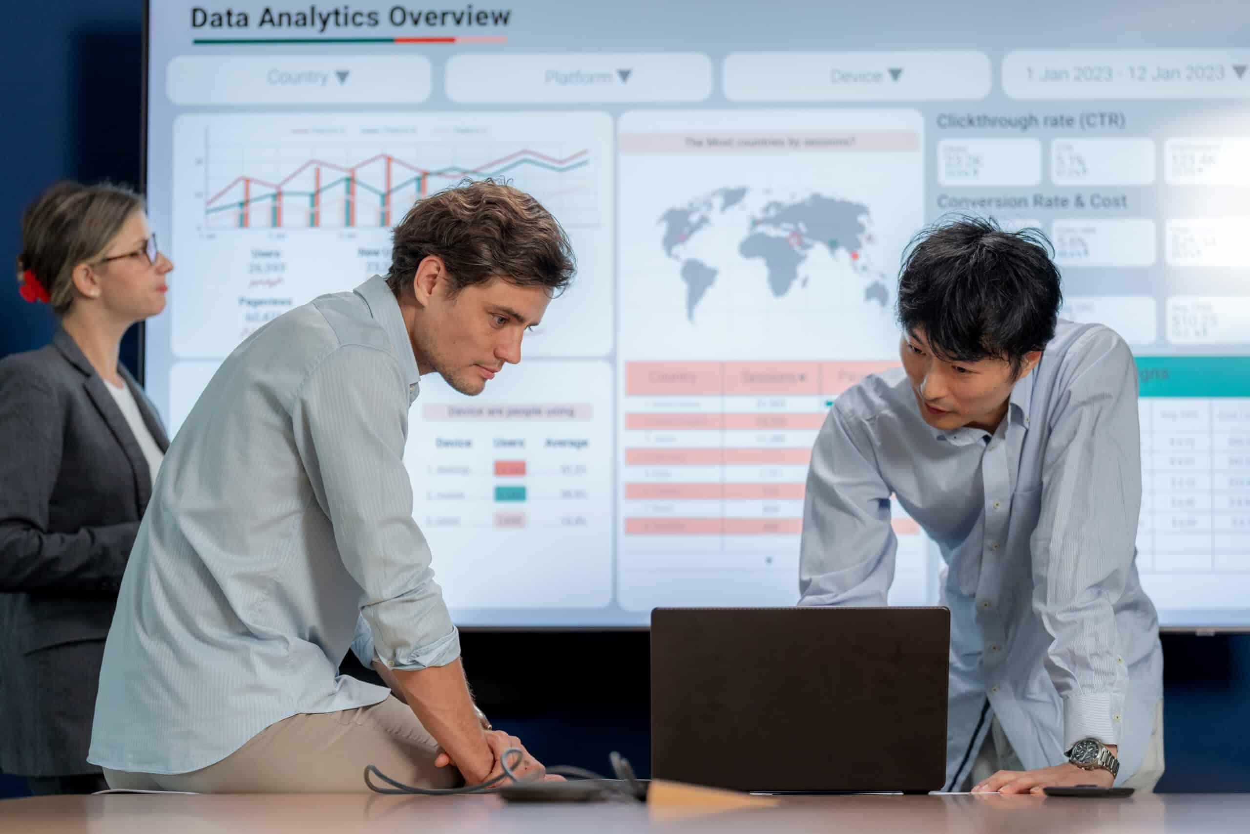 Two men discuss data at a laptop with analytics charts on a large screen behind them.