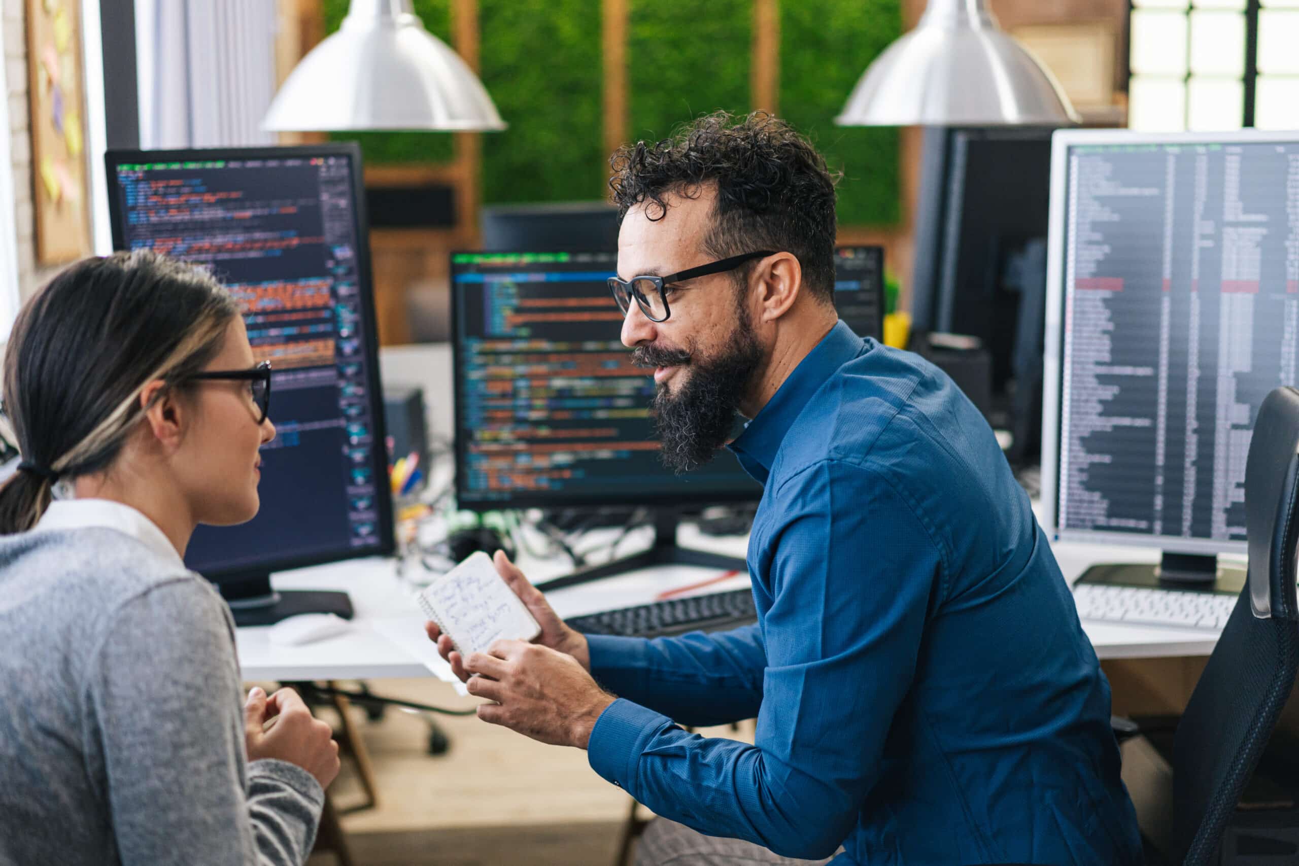 Two coworkers discuss Enterprise Systems Transformation by code on multiple office monitors.