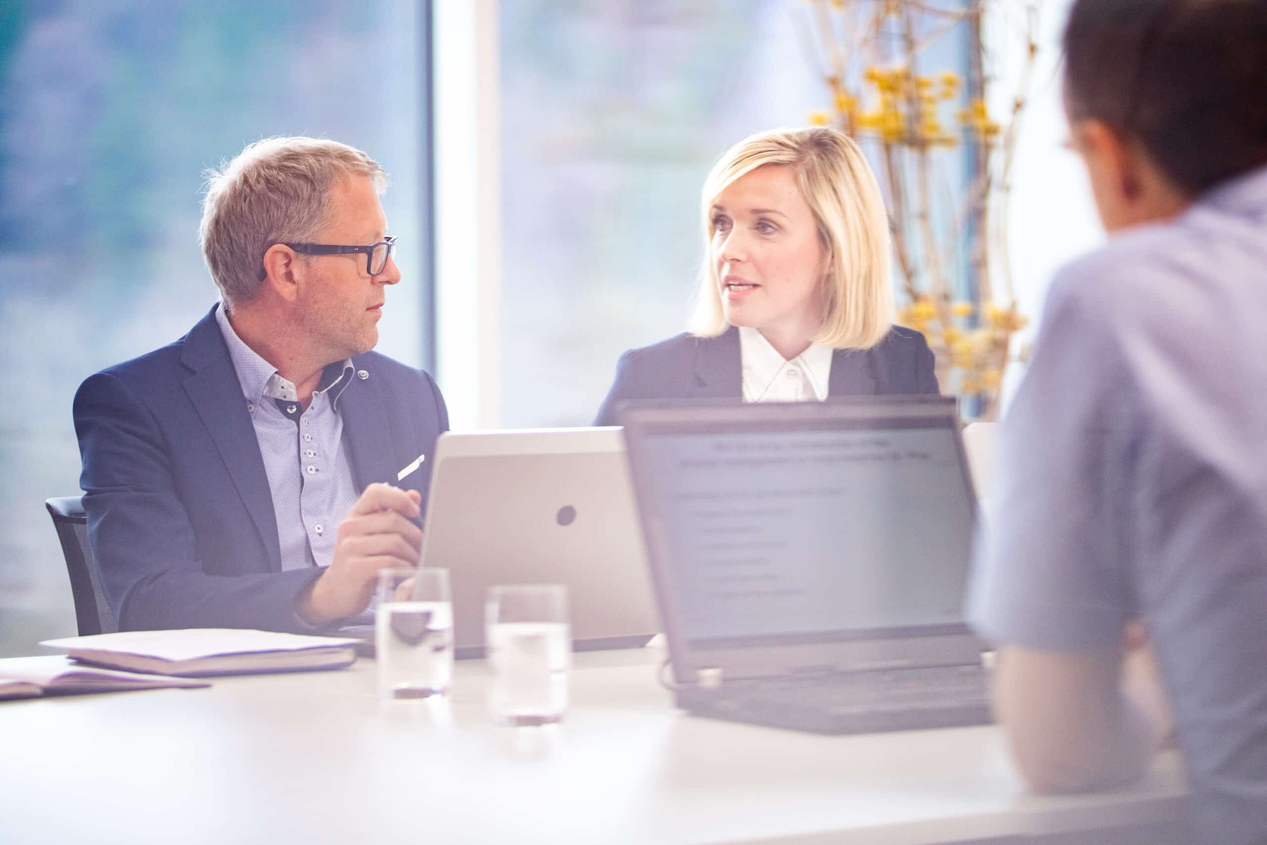 Three people in a meeting discuss Geospatial Analytics with laptops and documents on a bright table.