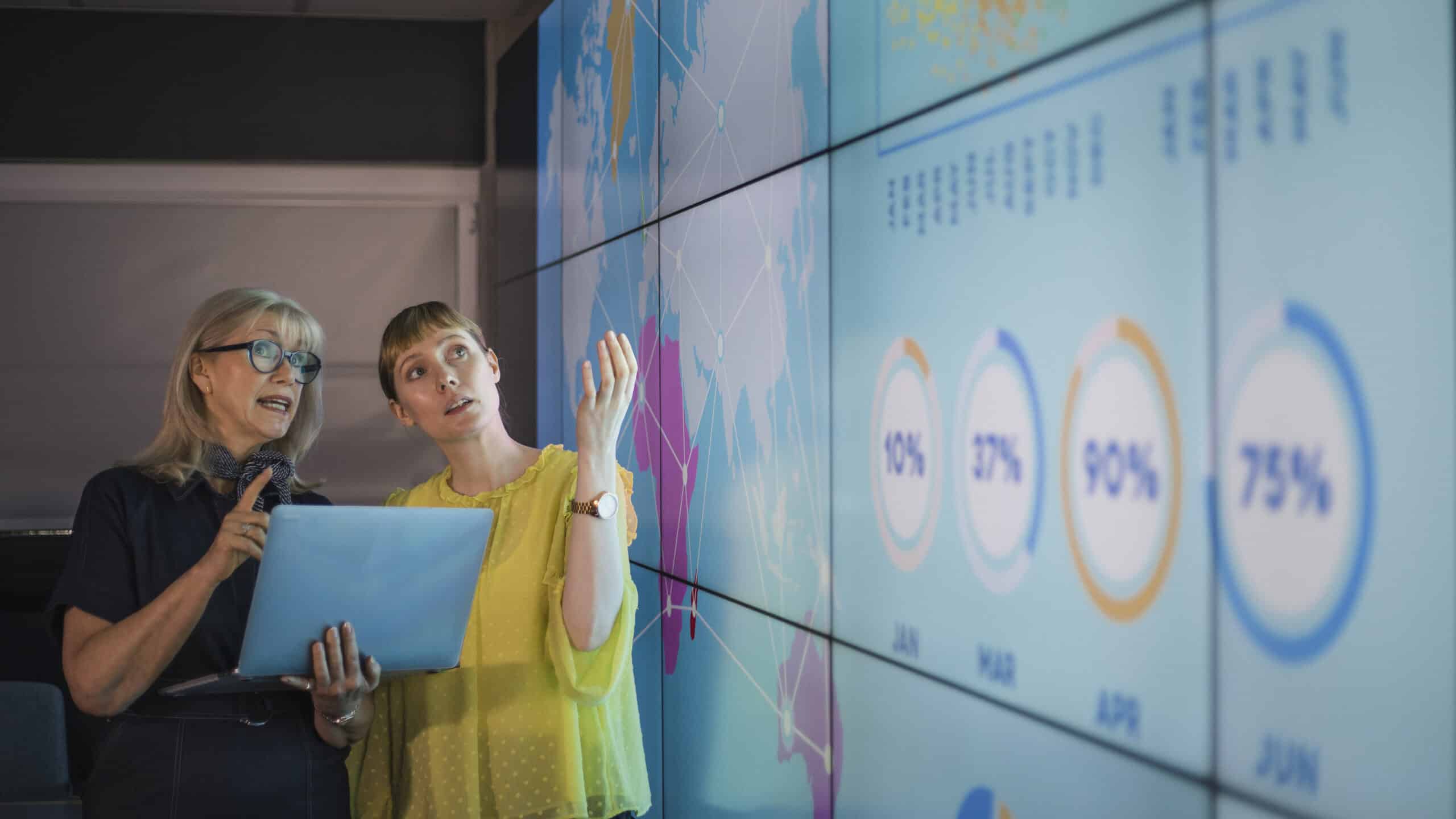 Two women discuss data on a large digital screen during an IoT consulting session.