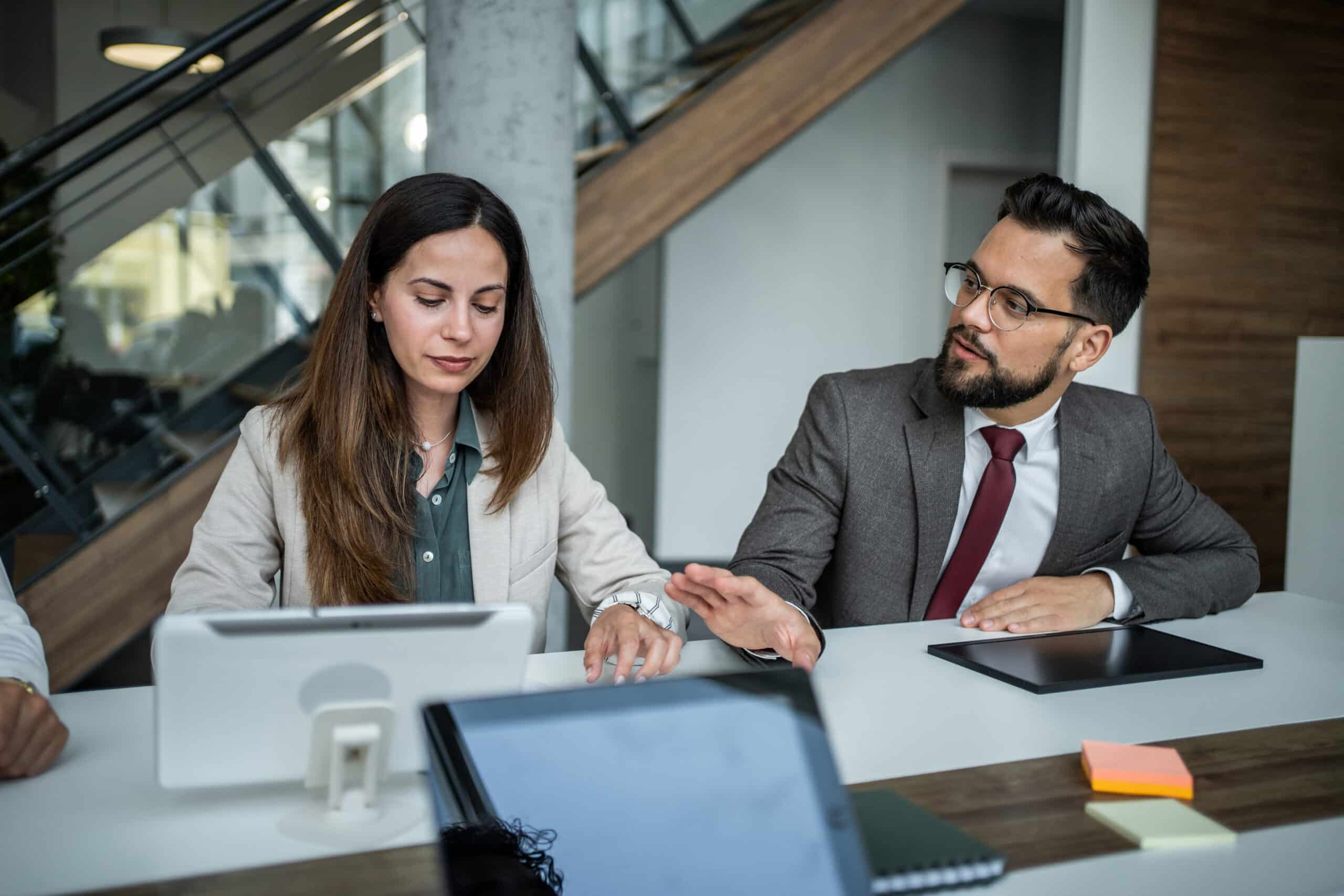 Two colleagues from Proptech Consulting discuss ideas at a modern office desk with tablets and notebooks.