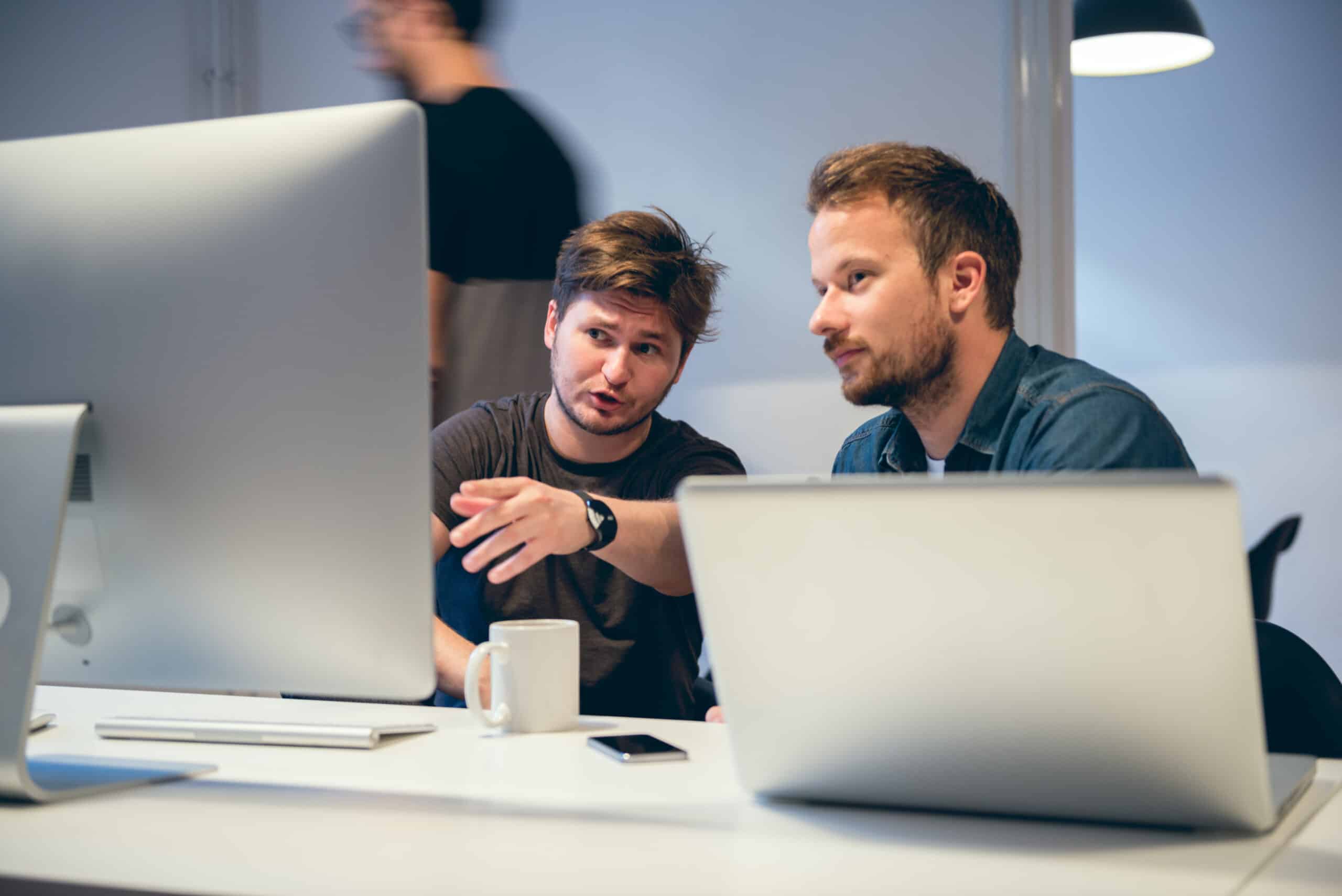 Two men, likely Demand Forecasting Consultants, discuss data on laptops over coffee at a desk.