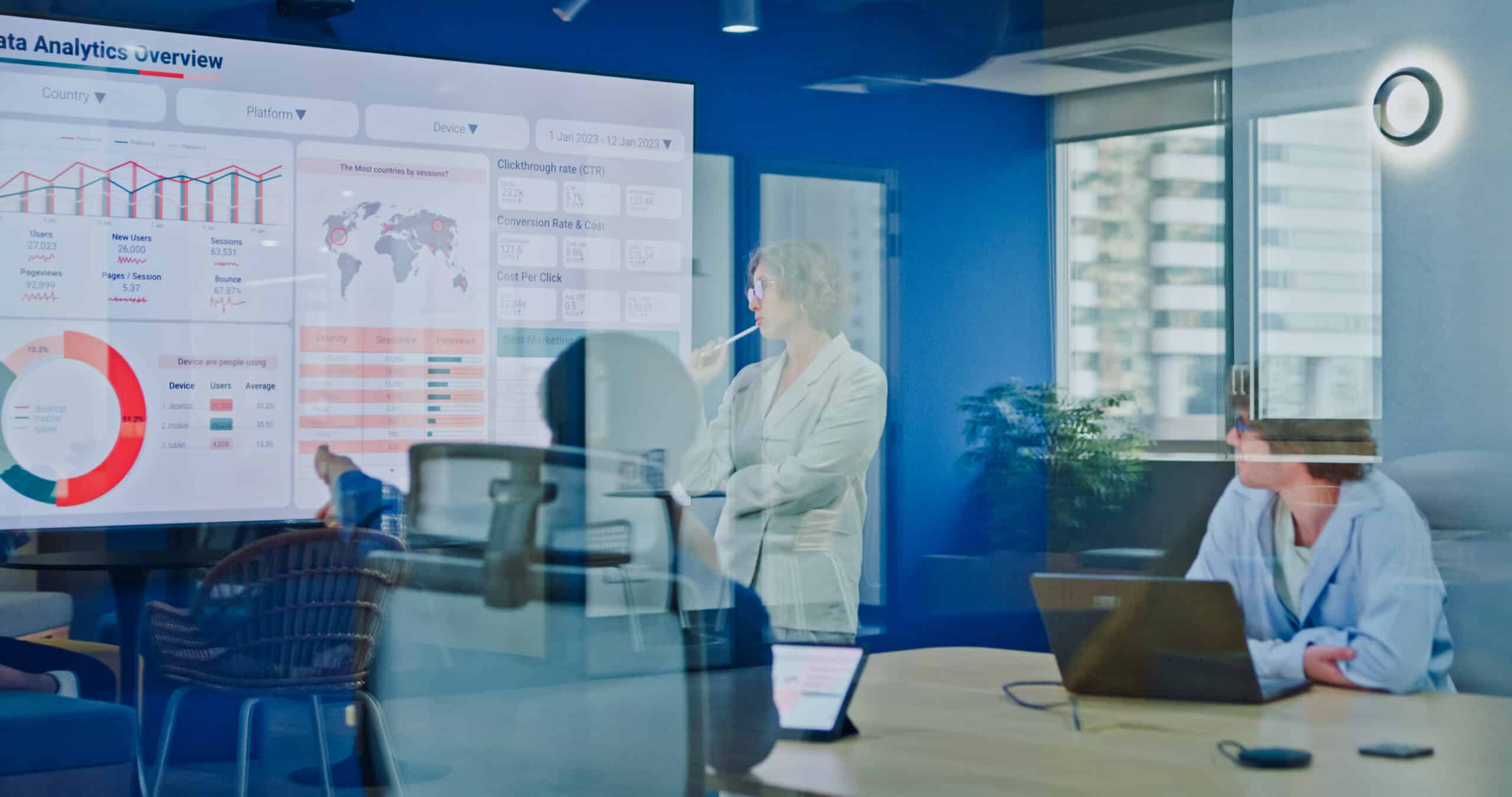 Two people in a meeting room discuss data analytics shown on a large screen.