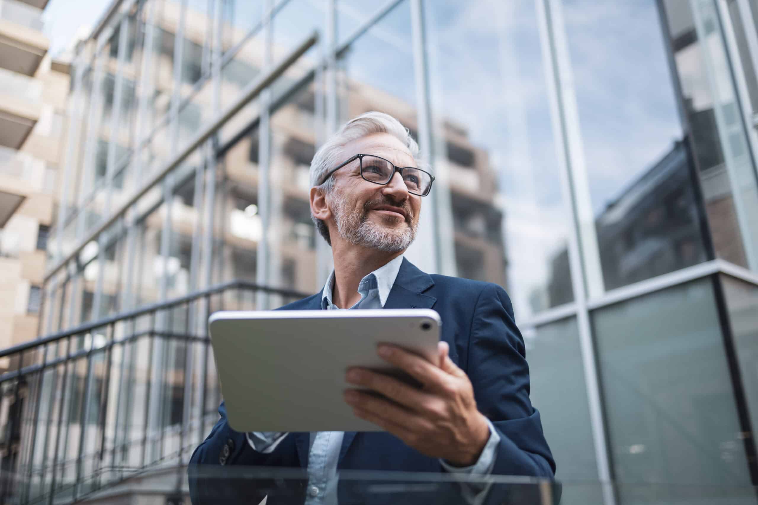 Smiling man in a suit holding a tablet outside a modern glass building.