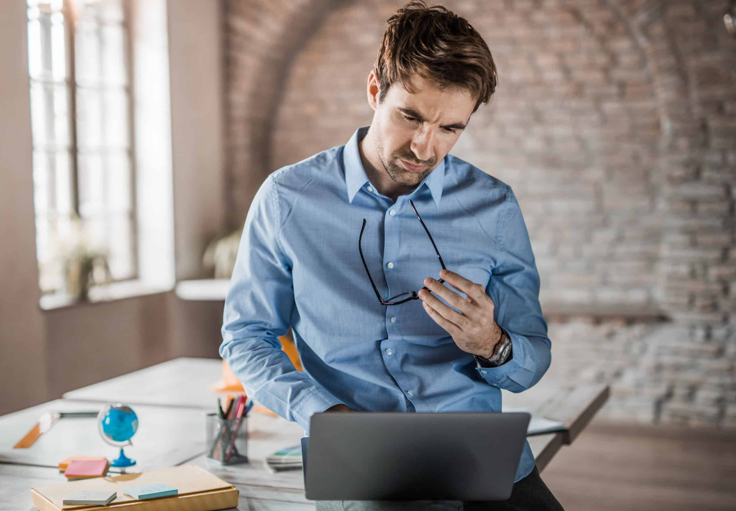 Man in blue shirt reviews IT Infrastructure Consulting plans on laptop, holding glasses at desk.