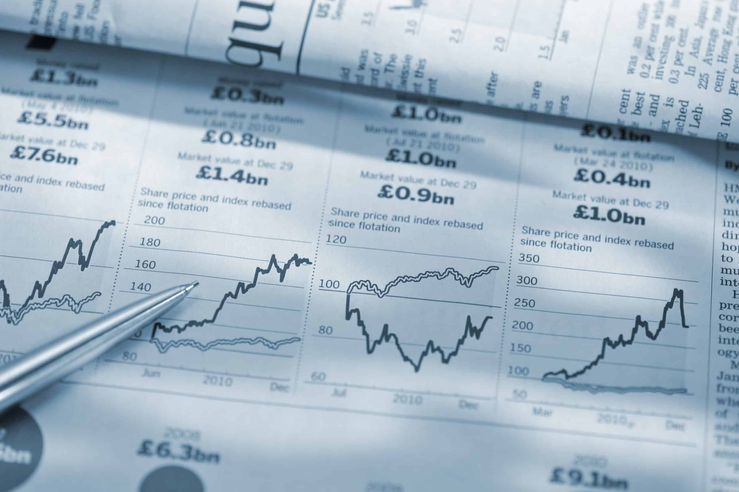 Pen pointing to line graphs and financial data printed on a newspaper page.