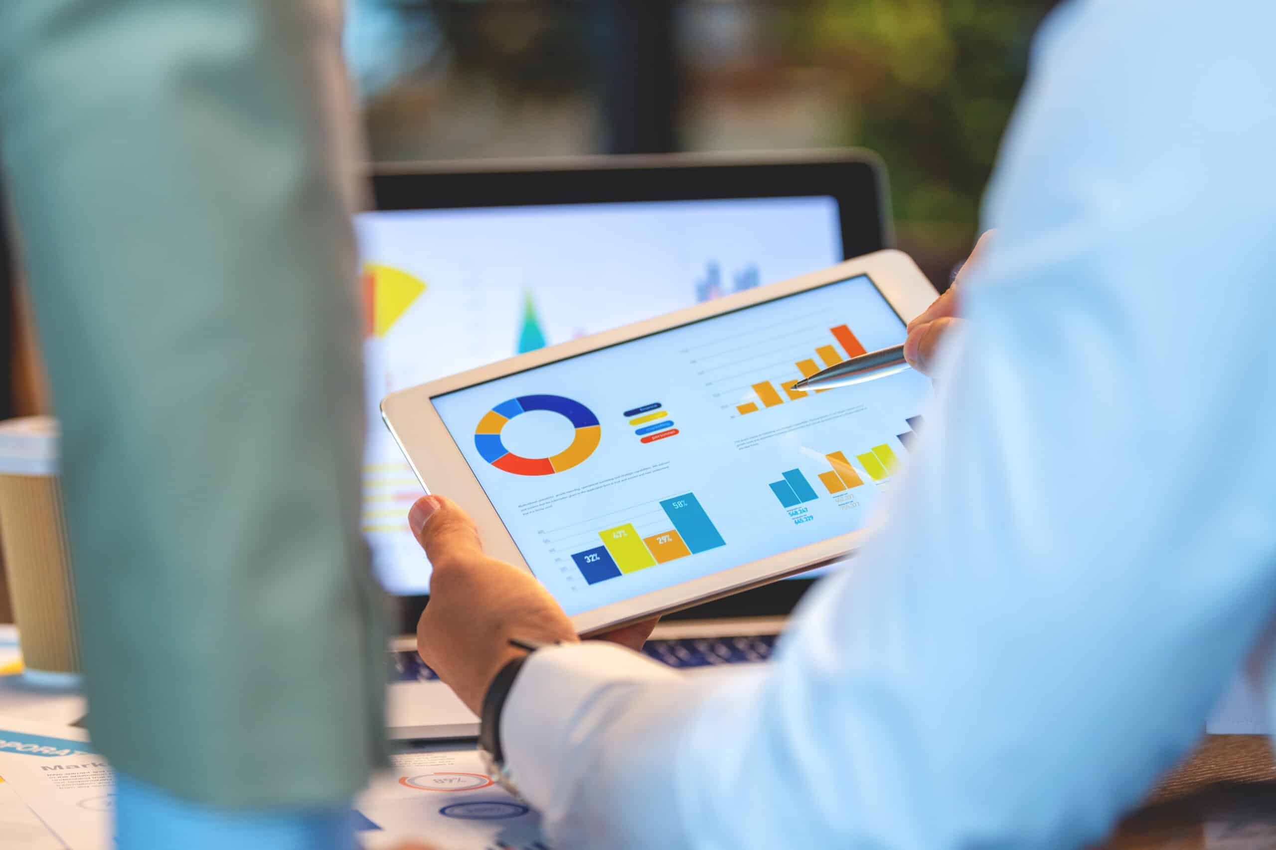 Person holding tablet displaying colorful charts and graphs, with laptop in background.