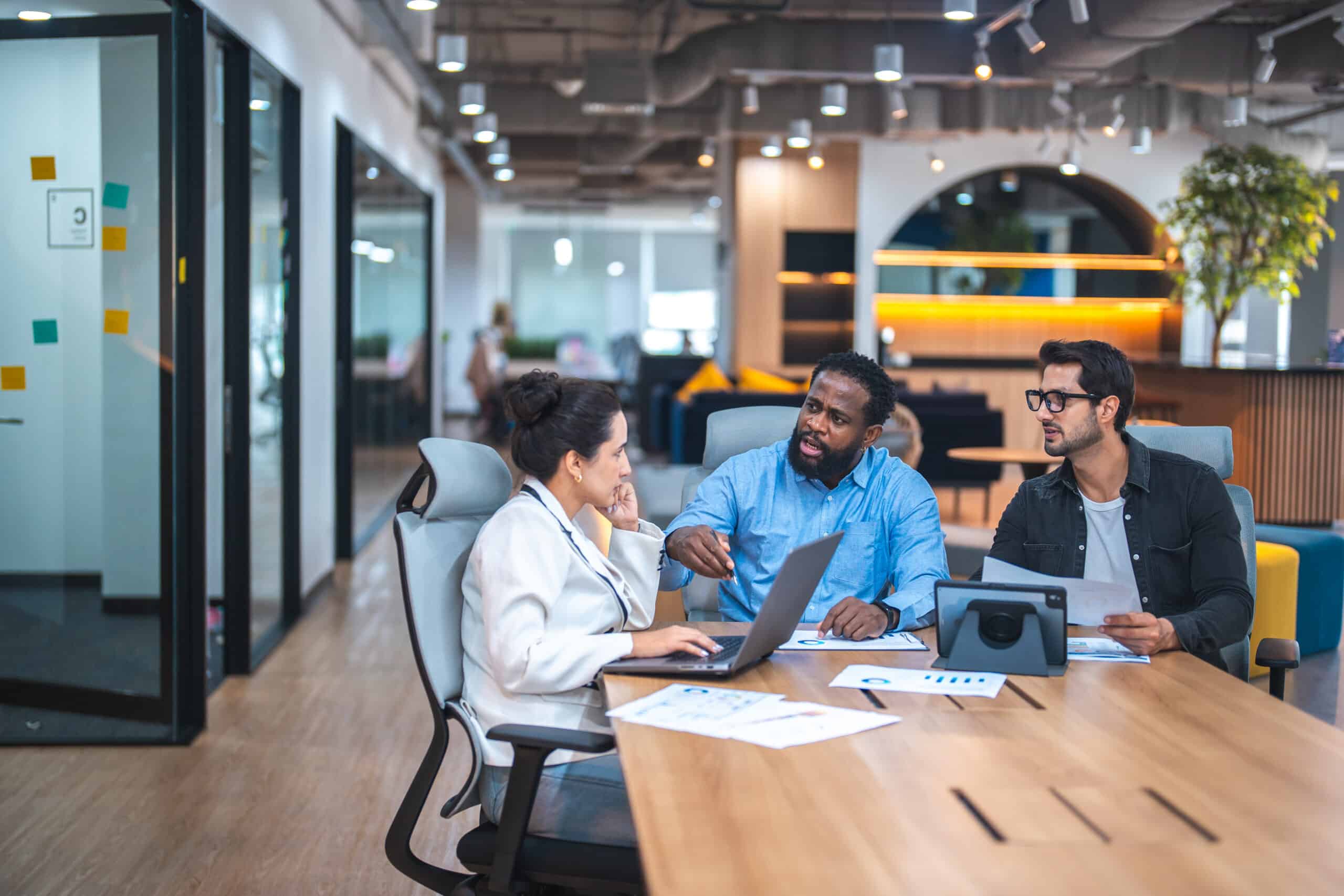 Three people discuss CRE Expense Reduction around a table with laptops in a modern office setting.