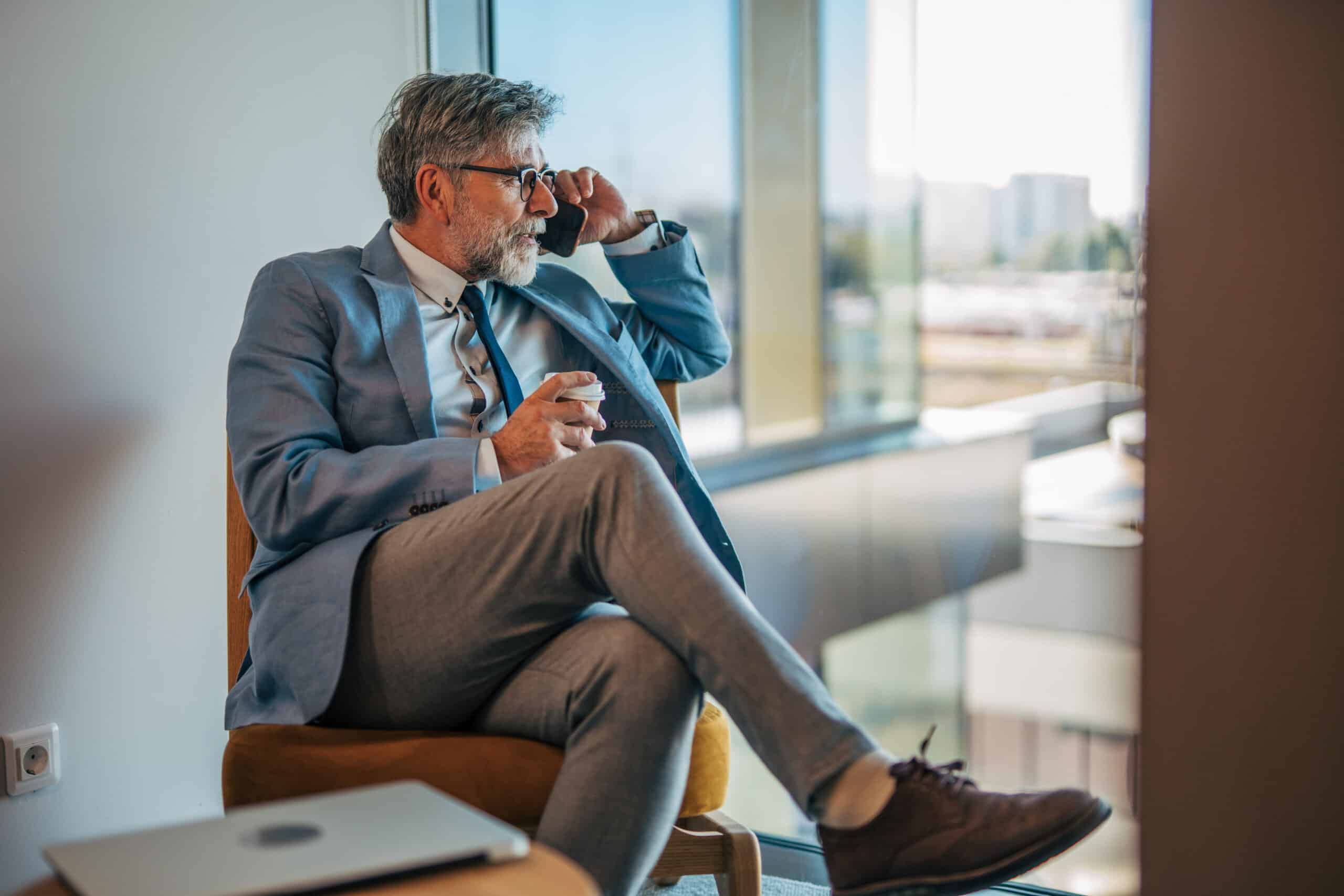 Man in a suit by the window discusses CRE Transaction Management over the phone, cup in hand.