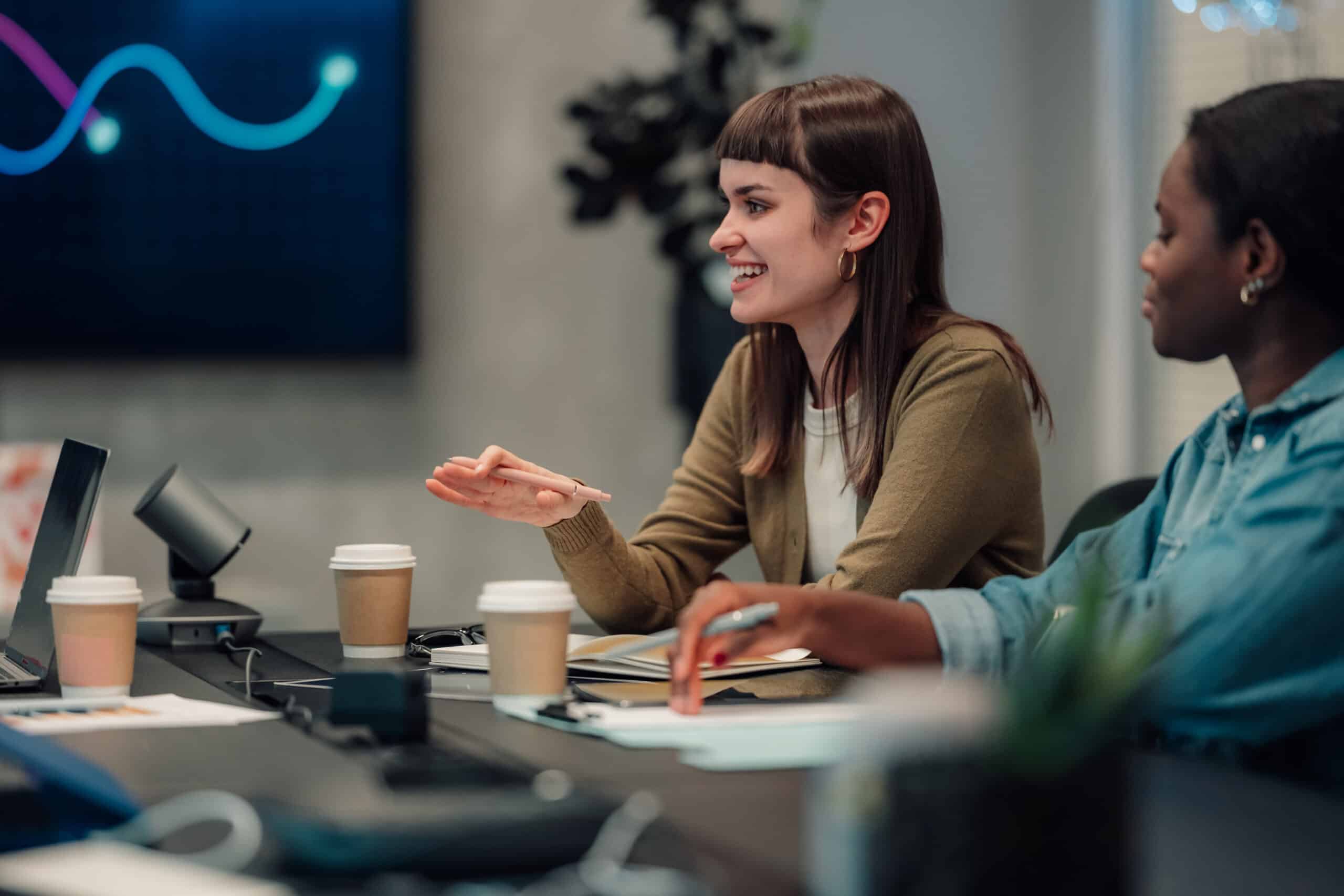 Two women smiling and talking at a Digital Twins Consulting meeting with laptops and coffee cups.