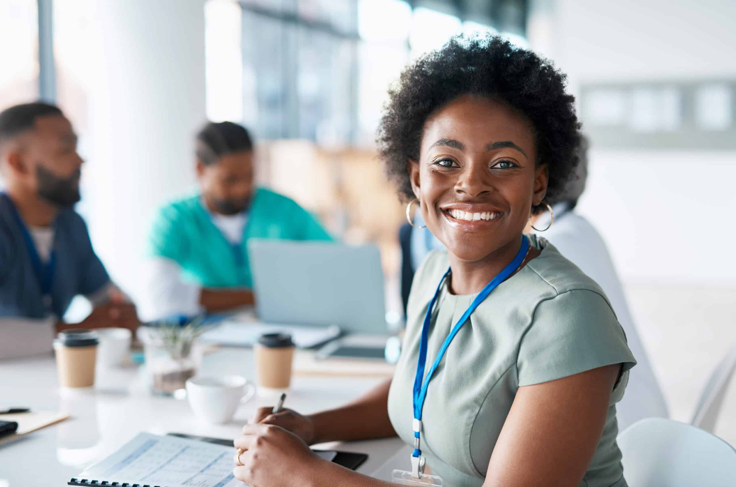 Smiling woman with a lanyard sits at a table with colleagues in a bright office.