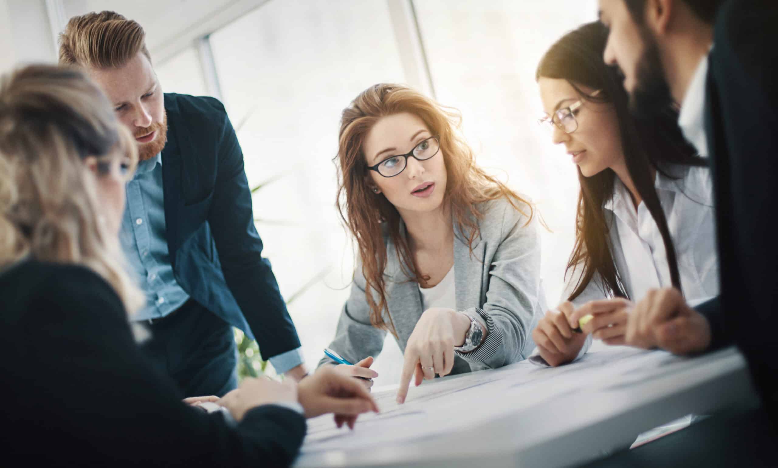 Five people in business attire discuss Leadership Development at a table in a bright office setting.