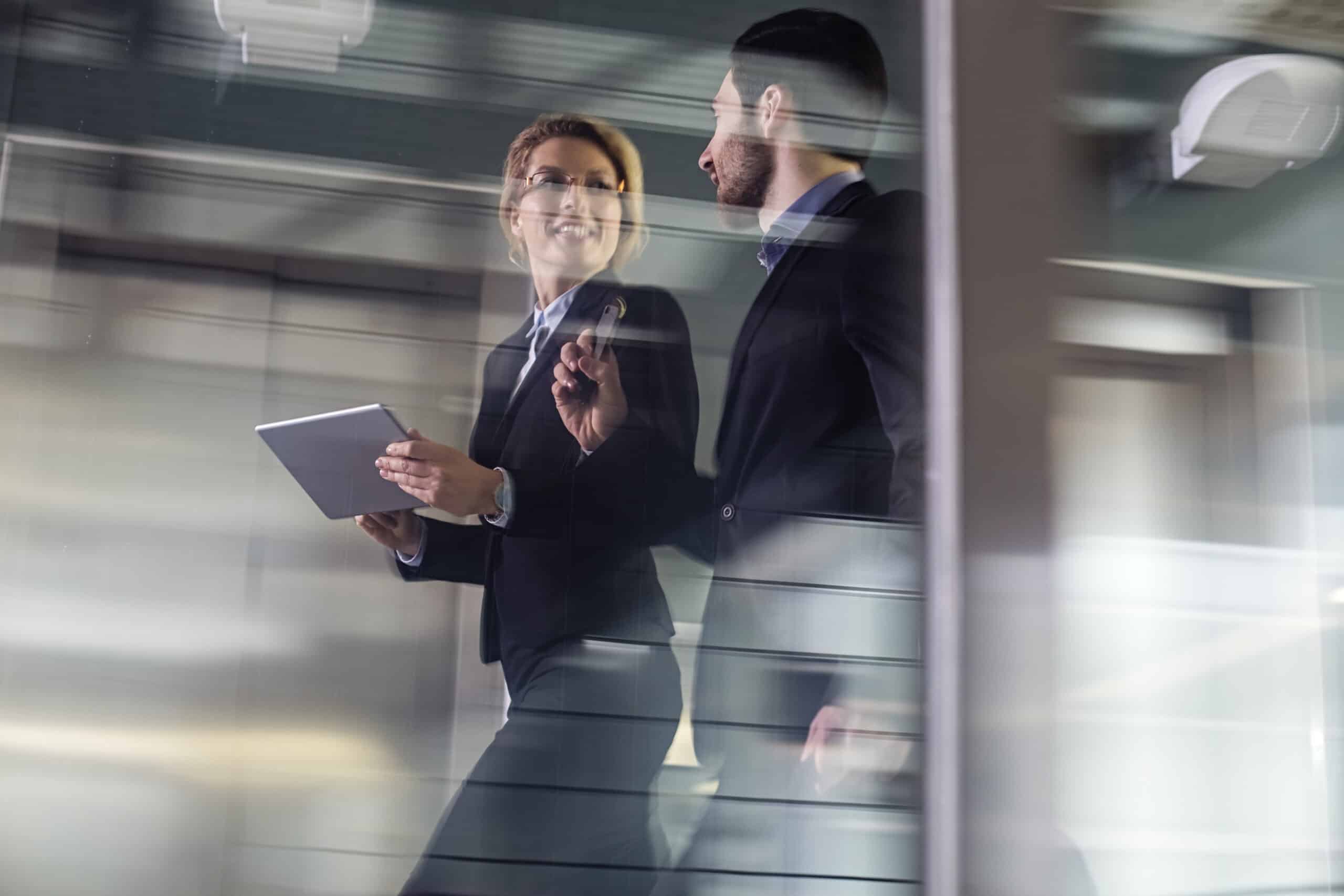 Two business professionals walking and talking inside an office building, one holding a tablet.