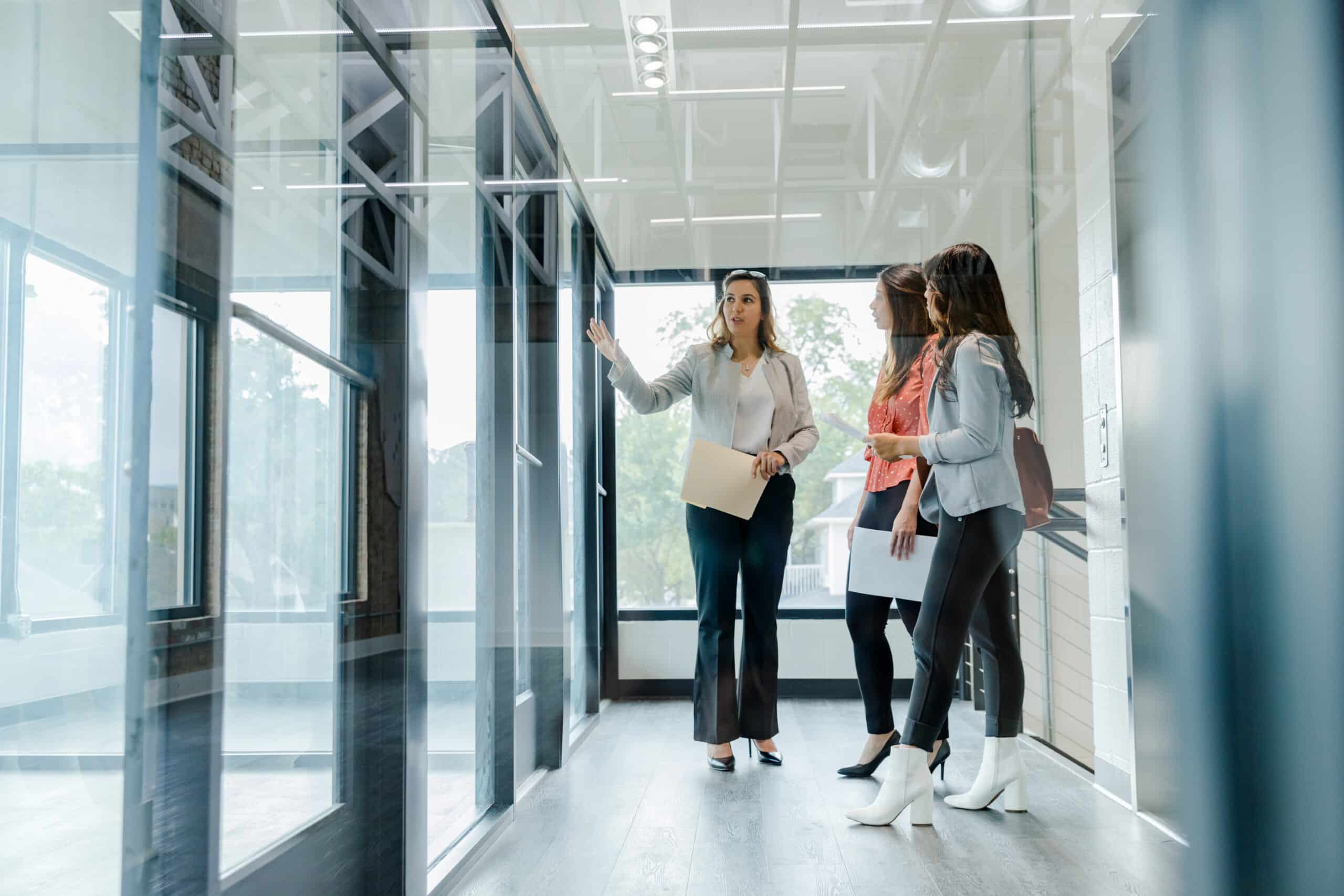 Three women tour a modern office space with glass walls and natural light.