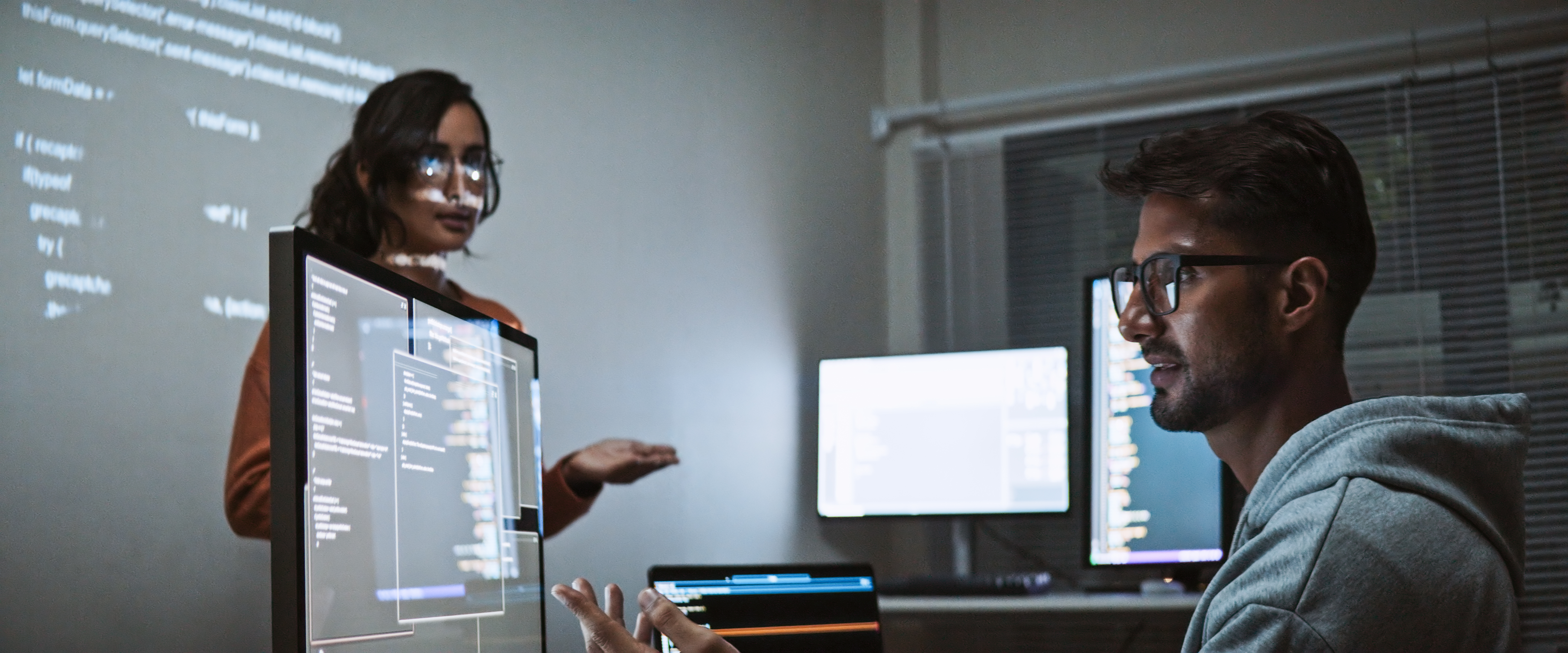 Two people discuss code and Intelligent Automation in a dimly lit office with code projections.