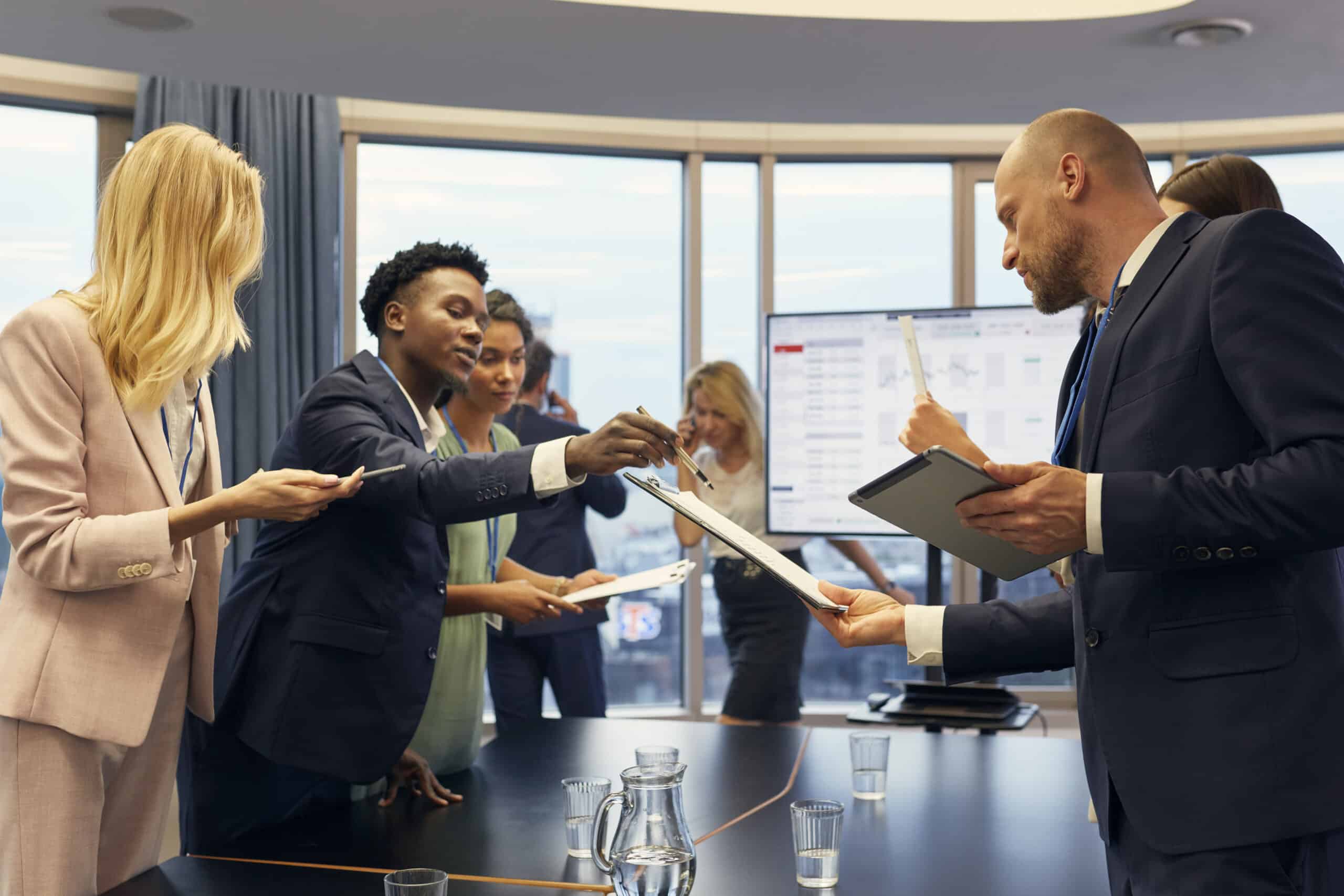 Businesspeople in a meeting exchanging documents around a conference table with digital devices.