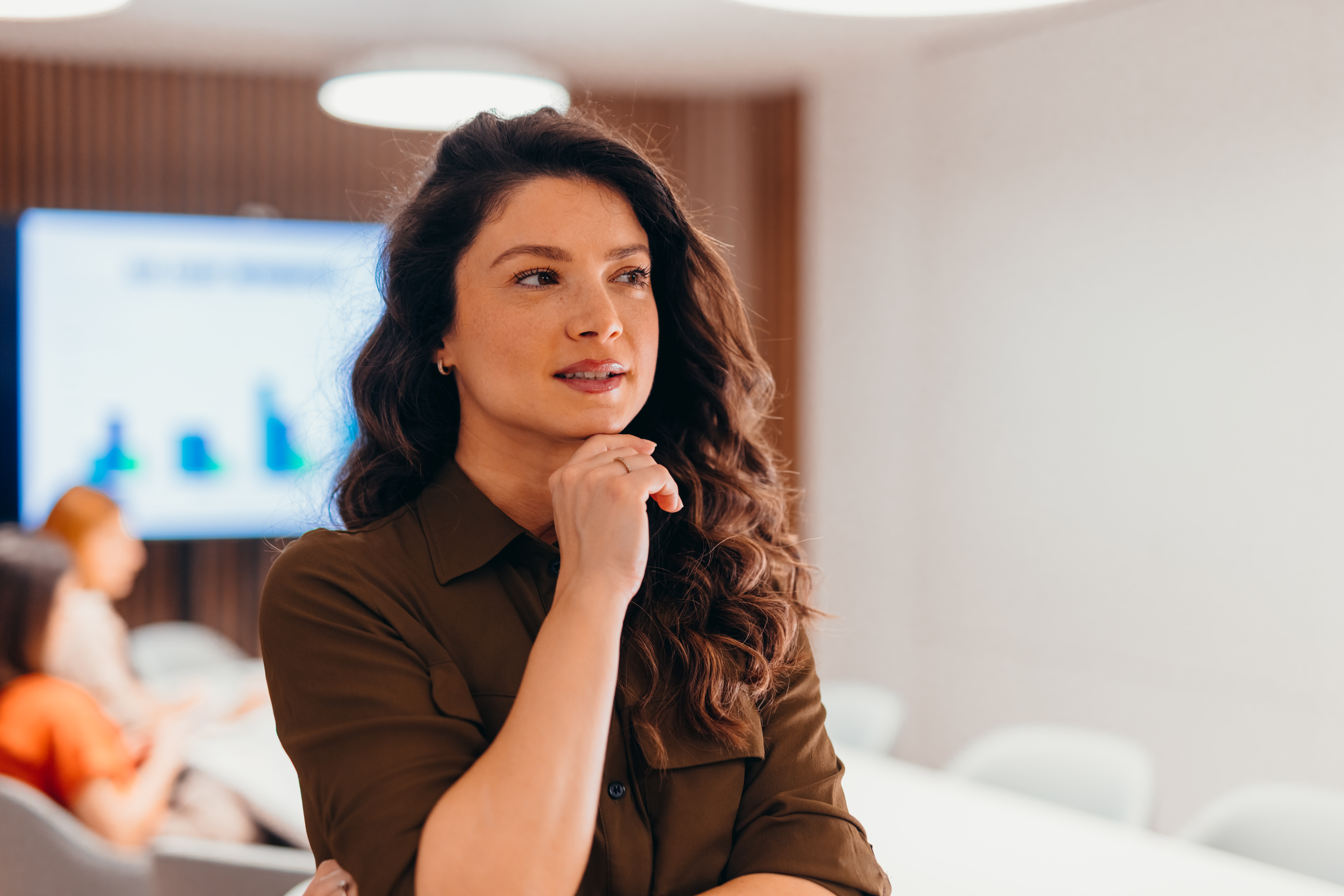 Woman with long hair thinking in a conference room, people and screen in background.