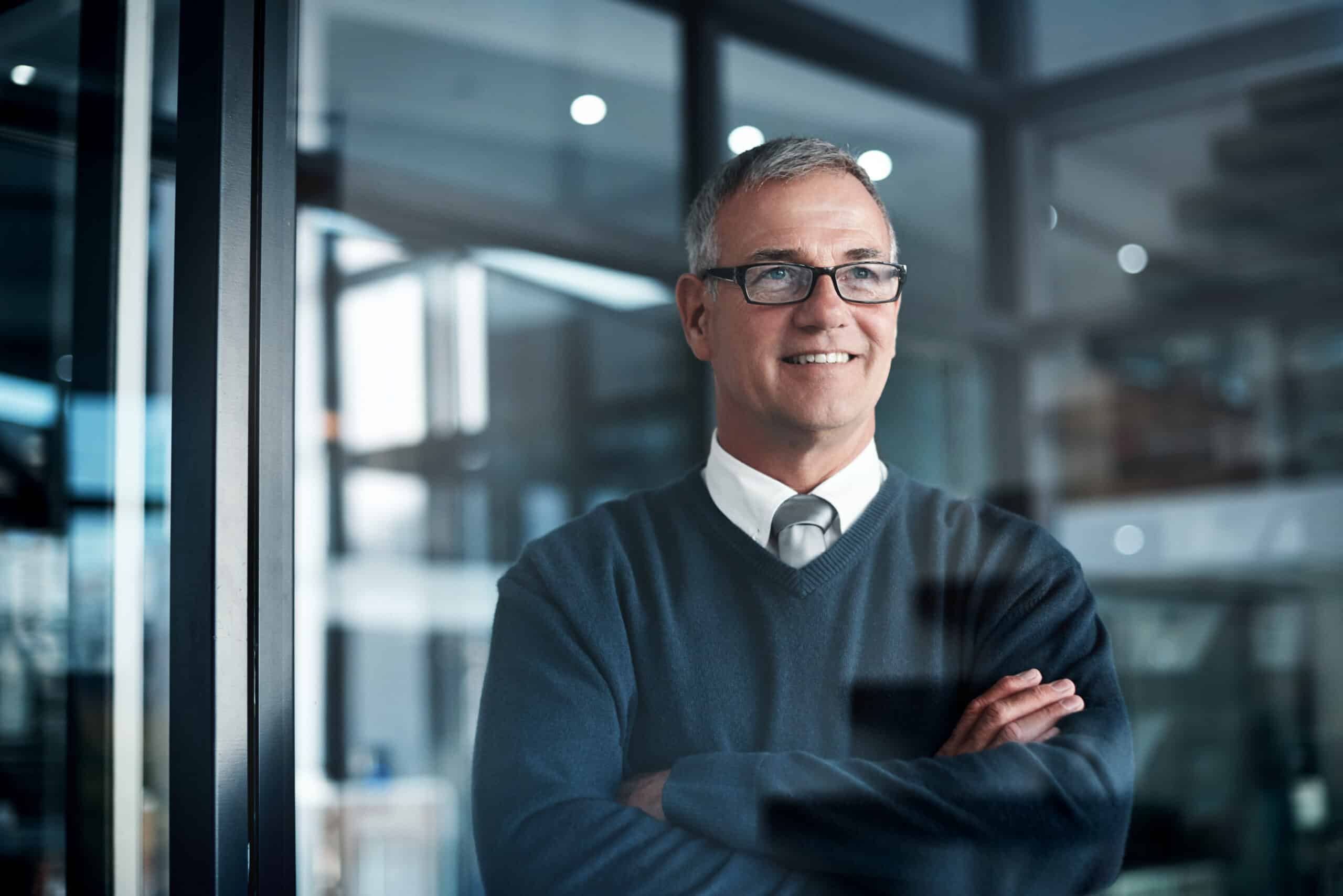 Smiling man with glasses and gray hair stands with arms crossed in an IT infrastructure office.