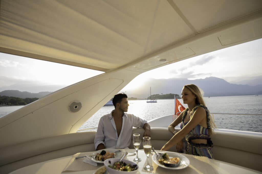 A man and woman enjoy a meal and drinks on a yacht with ocean and mountains in the background.