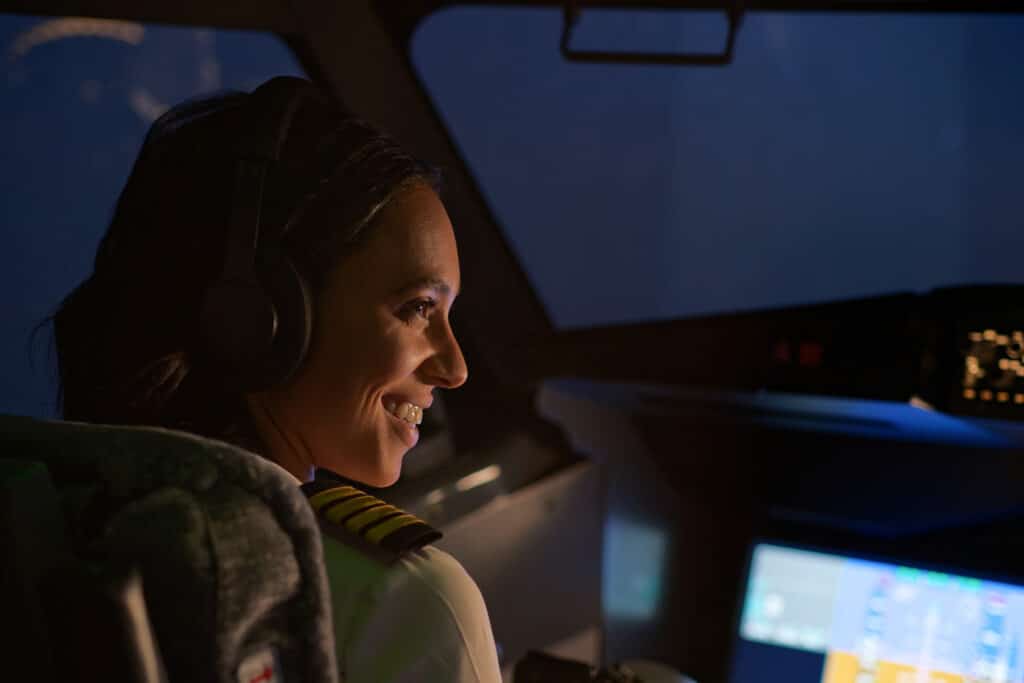 Smiling pilot wearing headphones sits in a dimly lit airplane cockpit.