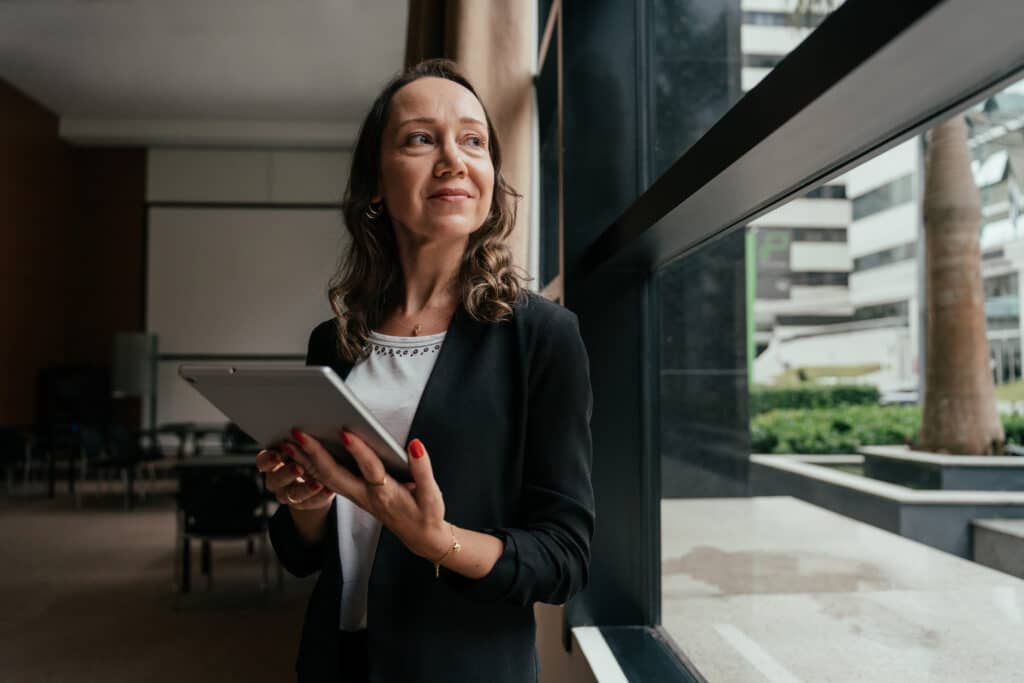 Woman in business attire with tablet, reflecting on executive search consulting by the window.