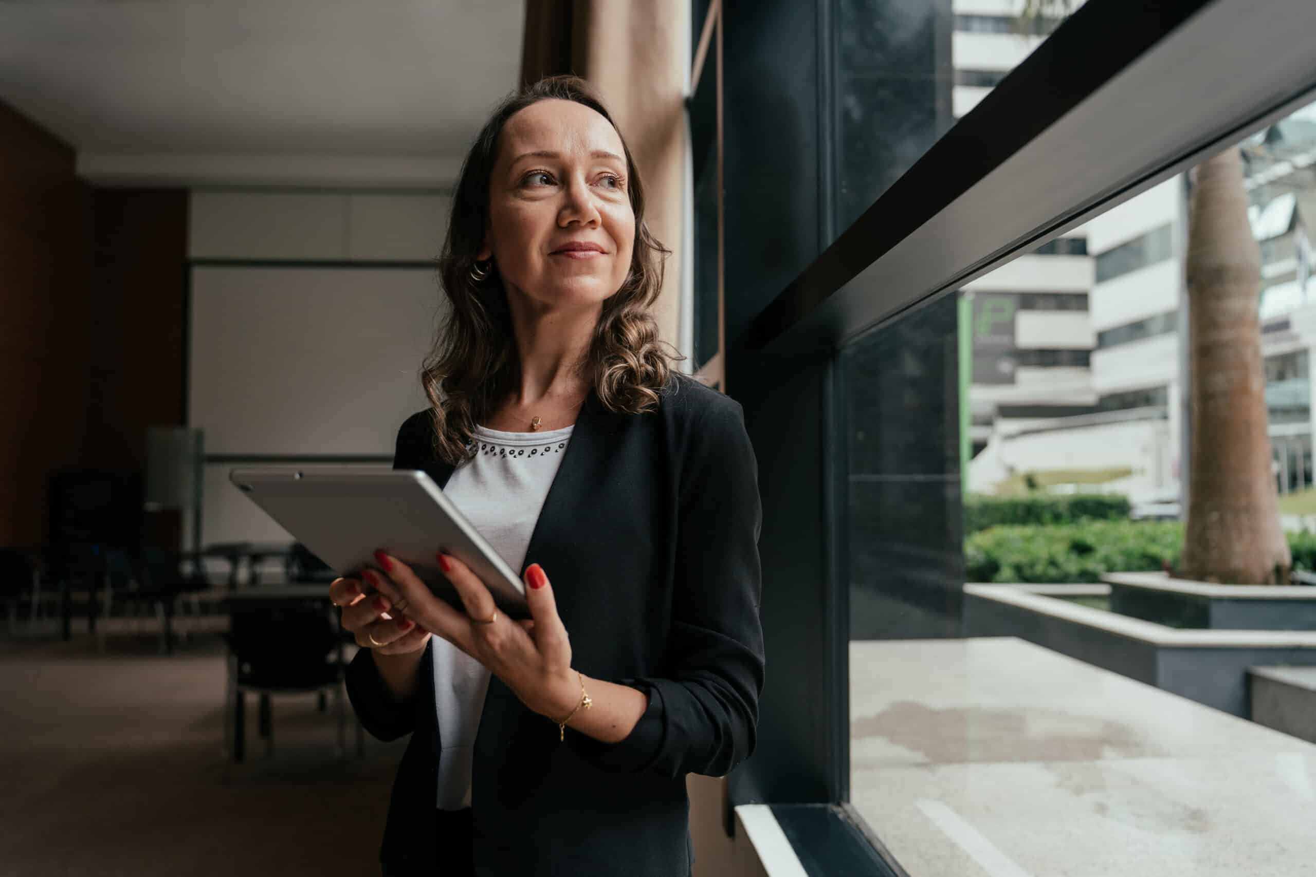 Woman in business attire with tablet, reflecting on executive search consulting by the window.