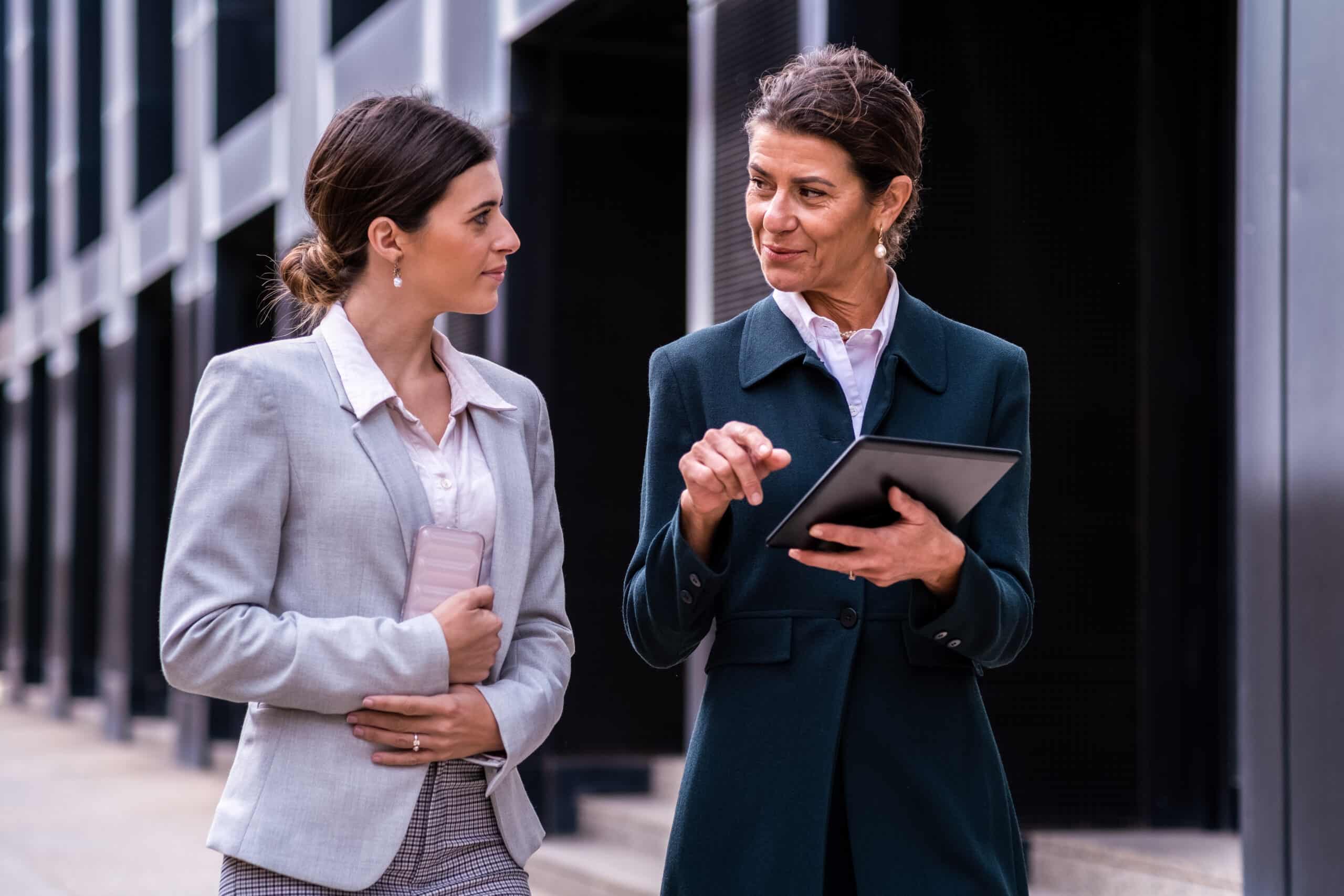 Two businesswomen discuss succession planning consulting outside, with a tablet and phone in hand.