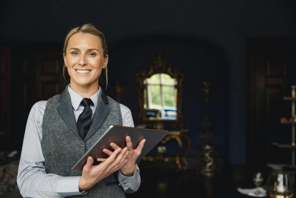 Smiling woman in a vest and tie holding a tablet in an elegant, dark room.