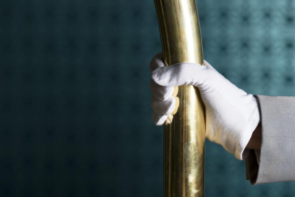A gloved hand holds a shiny brass pole, reflecting luxury hospitality, against a teal background.