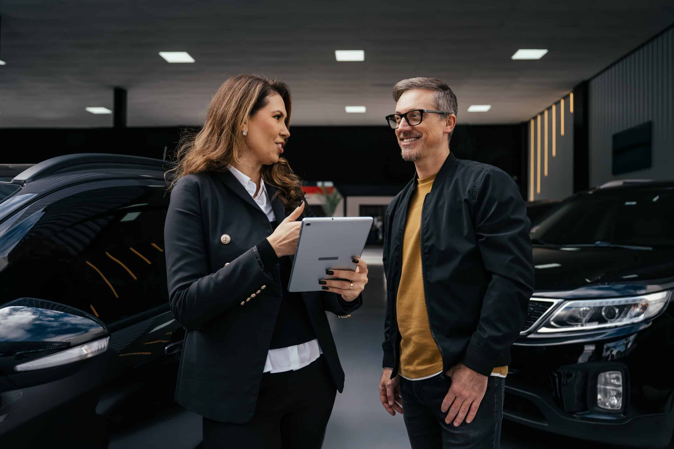 Woman shows tablet to man, enhancing automotive customer experience consulting near black vehicles.