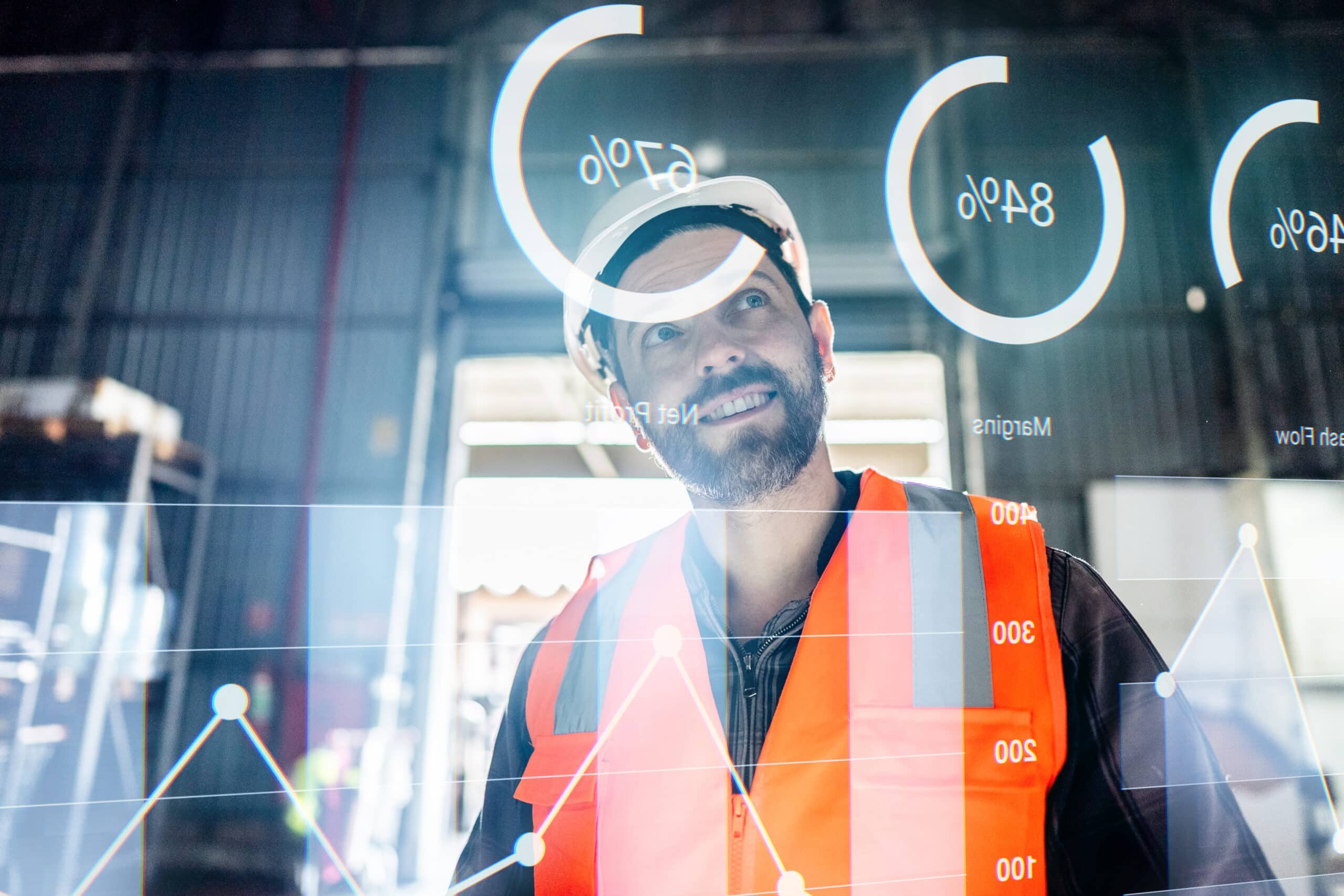 Man in a hard hat and vest reviews industrial IoT consulting graphs in a warehouse.