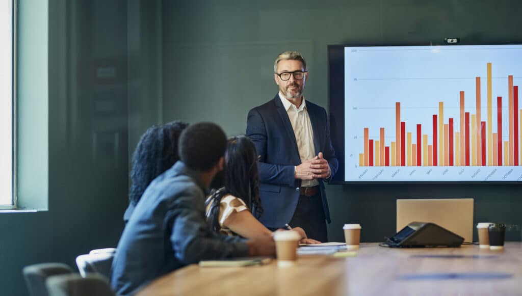 Man presenting bar graph to colleagues in a Revenue Management Consulting conference room.