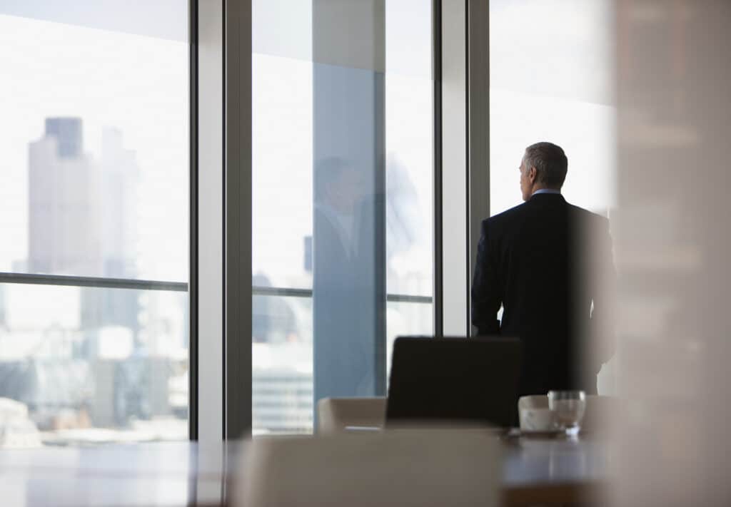 Man in suit looks out office window, pondering how AI is reshaping counsel and city economics.