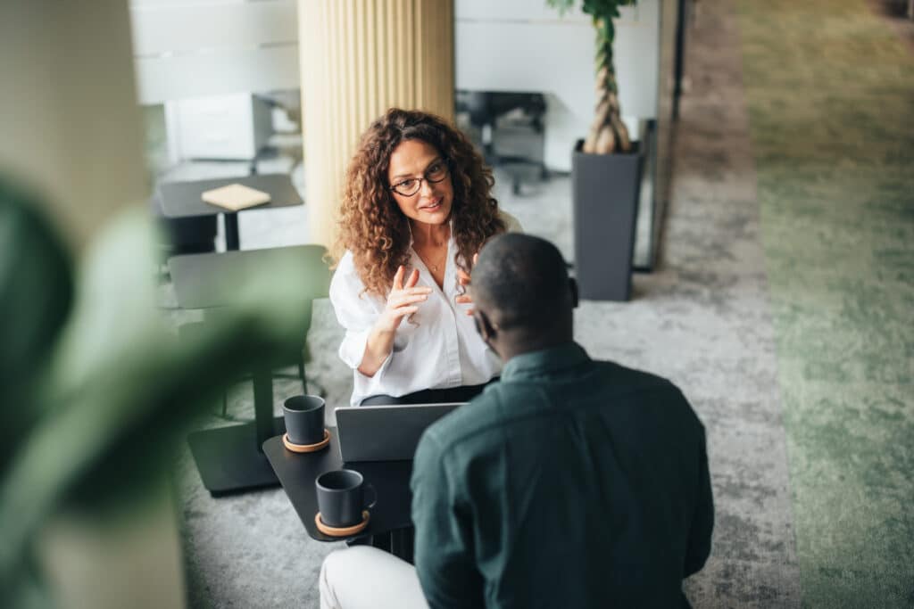 Two people discuss Designing Capacity for Flatter Organizations at a small table in a modern office.