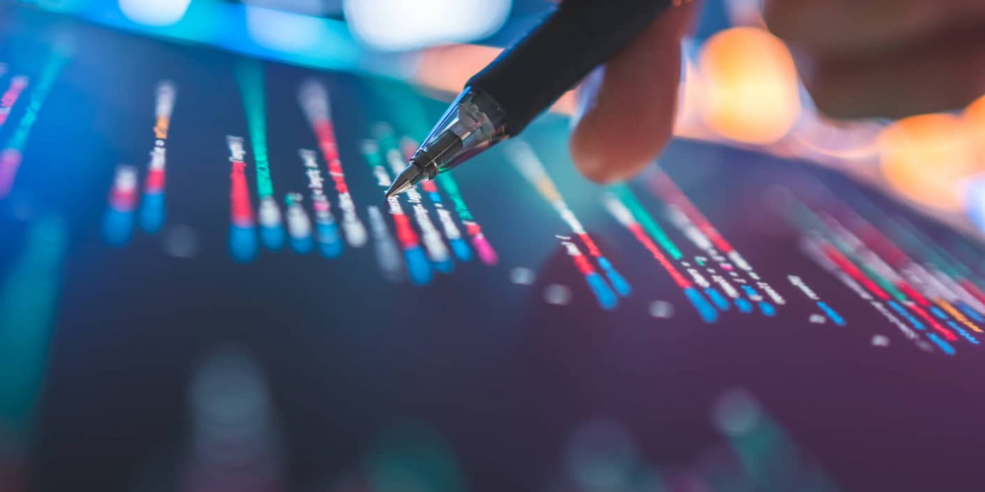 Hand holding pen points at colorful digital financial charts on a screen, analysis concept.